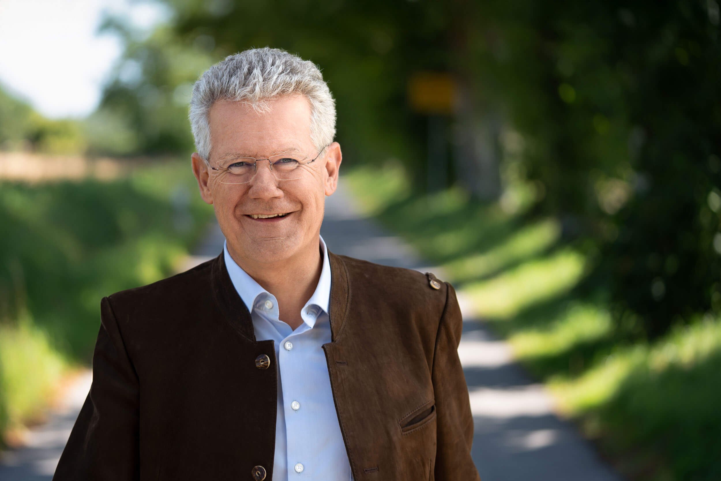 Älterer Mann mit Brille, grauem Haar und braunem Jacke, steht auf einer begrünten Straße, lächelt in die Kamera.