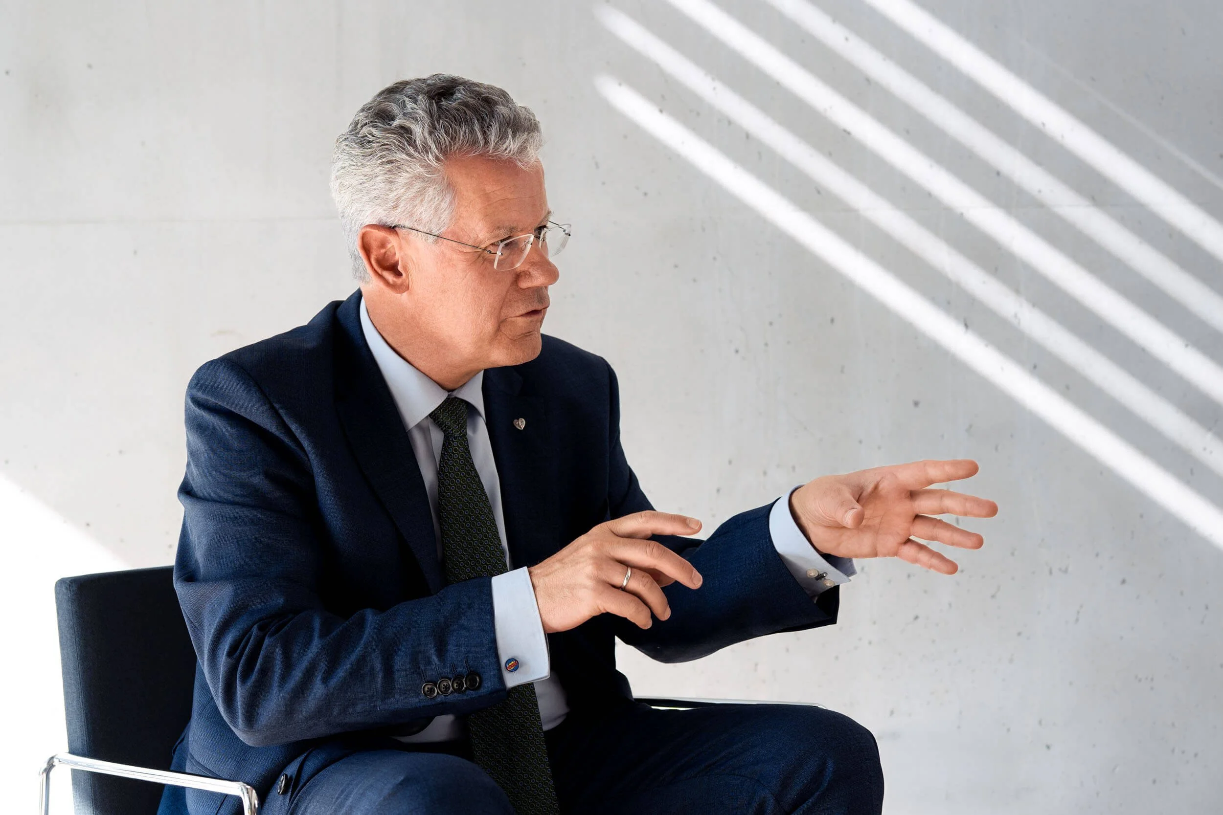 Ein älterer Mann in formeller Kleidung sitzt in einem weißen Raum mit modernem Design und gestreiften Schatten an der Wand. Er trägt eine dunkle Anzugjacke, ein weißes Hemd, eine dunkle Krawatte und eine Brille. Er ist in einer nachdenklichen Pose, wobei seine Hände in der Luft schweben und er den Blick nach unten gerichtet hat.