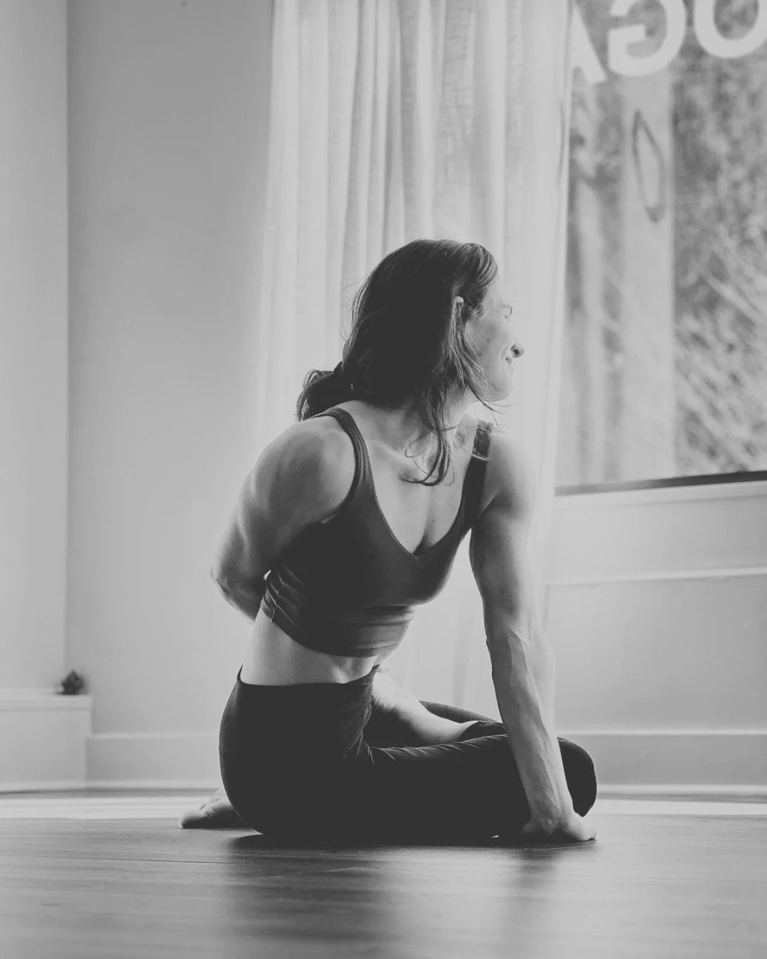 A woman practicing a posture from the second series of ashtanga yoga on a wooden floor near a large window with curtains, in black and white.