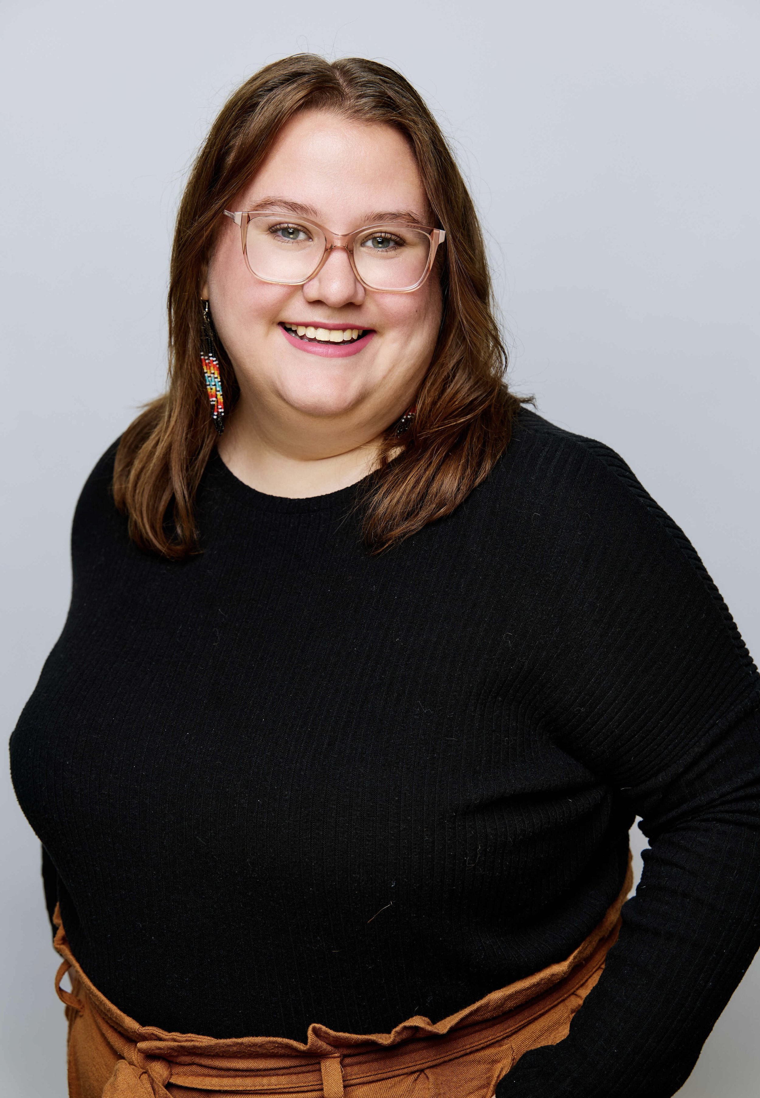 A smiling woman with shoulder-length brown hair and glasses, wearing a black top and orange-brown pants, posing against a light gray background.