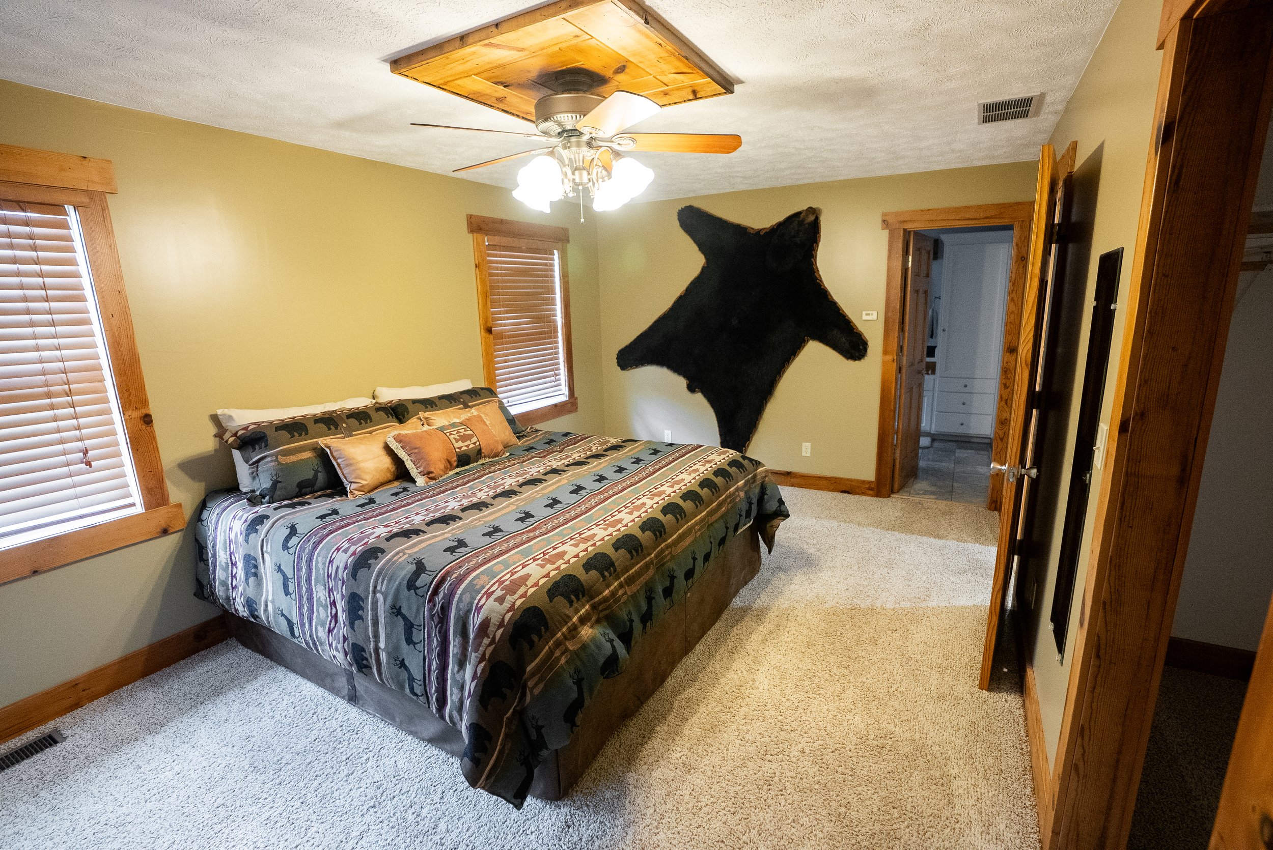 Bedroom with a bed featuring animal print bedding, wooden window frames, beige carpet, ceiling fan, and a large black bear rug hanging on the wall.