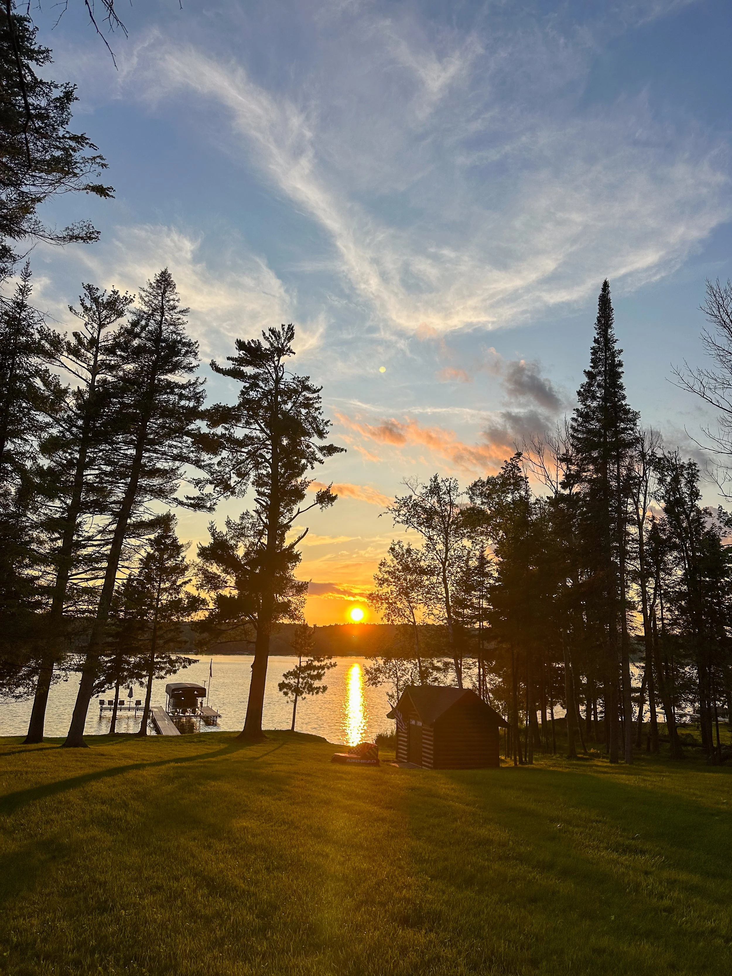 A sunset over a lake with reflected sunlight, surrounded by tall trees, a small cabin, and a dock with a boat.