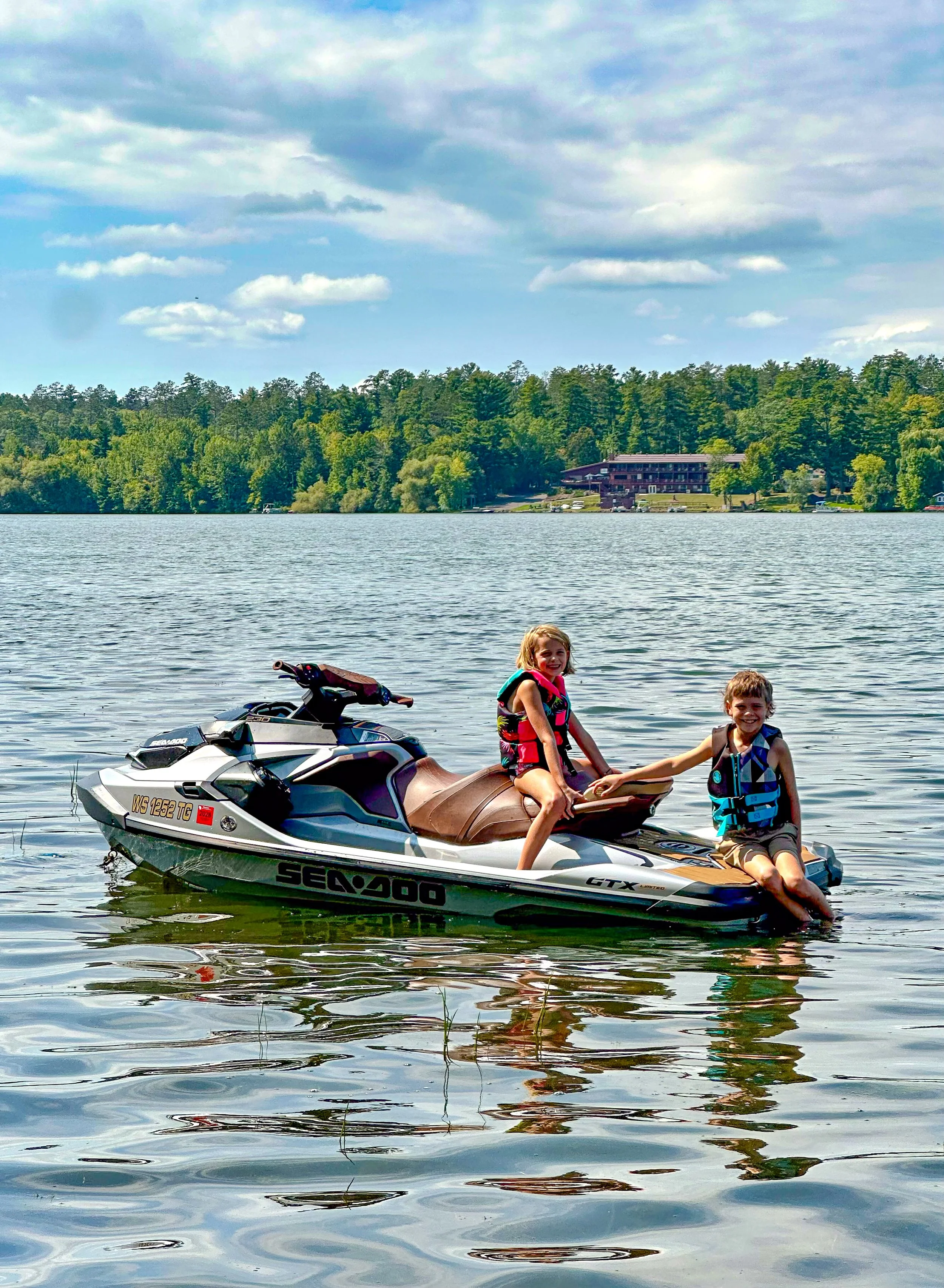 Two children on a Sea-Doo jet ski in a lake, wearing life jackets and smiling, with green trees and houses in the background under a partly cloudy sky.