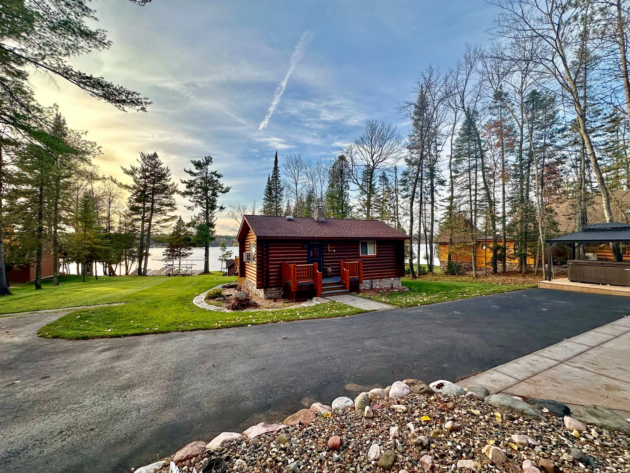 A small red cabin with a front porch sits on a grassy area next to a driveway. Trees surround the scene, and a lake is visible in the background with a partly cloudy sky overhead.