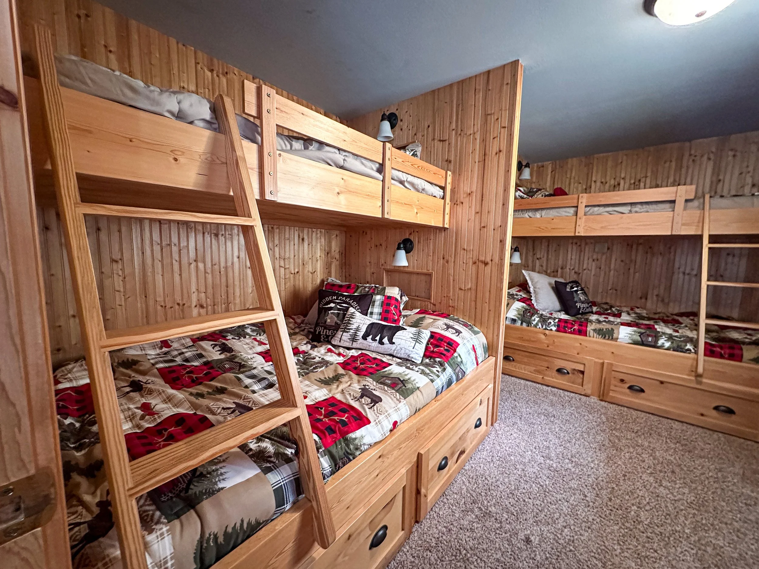 A room with three sets of bunk beds, all made of wood, each with pillows and patterned bedding with Christmas-themed designs. The beds have ladders for access, and the room has wooden walls, carpeted flooring, and wall-mounted lights above each bed.
