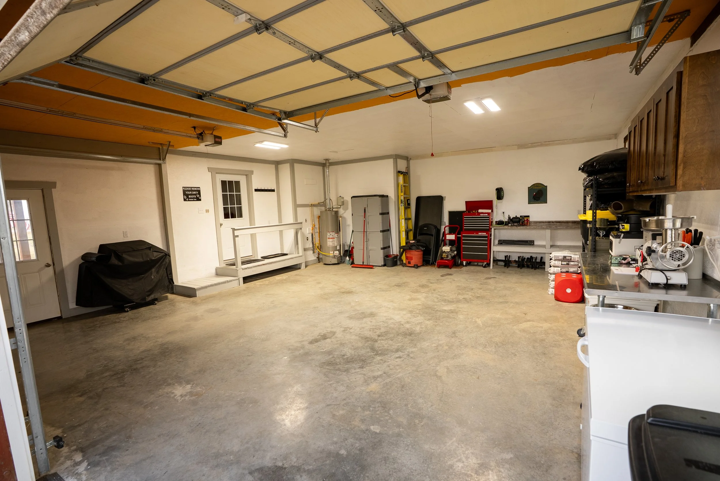 A spacious garage with a concrete floor, white walls, and various tools and storage units along the right wall. The garage door is open, revealing the ceiling mechanism.