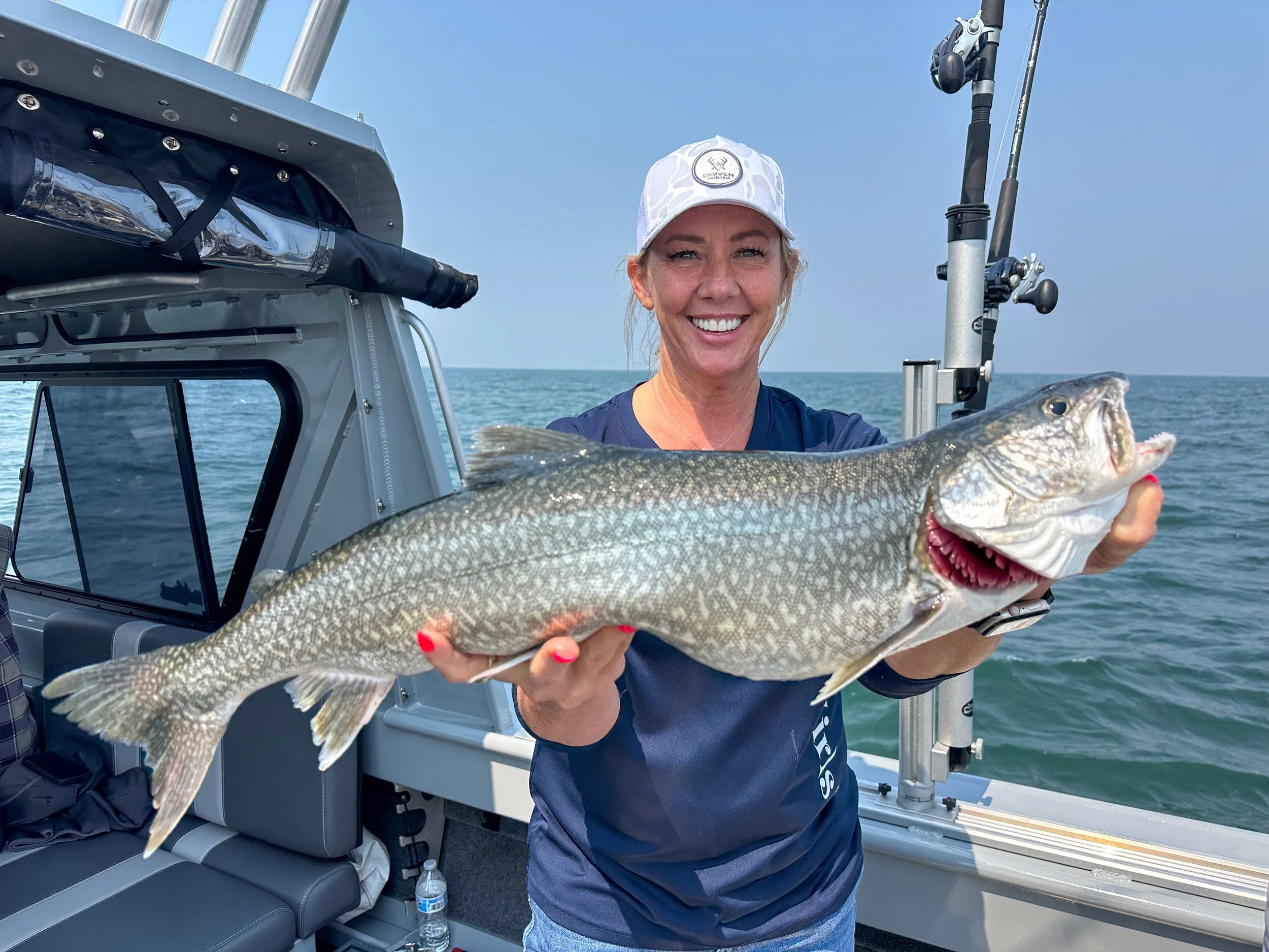 Woman smiling and holding a large fish on a boat in the ocean, with fishing gear visible nearby.