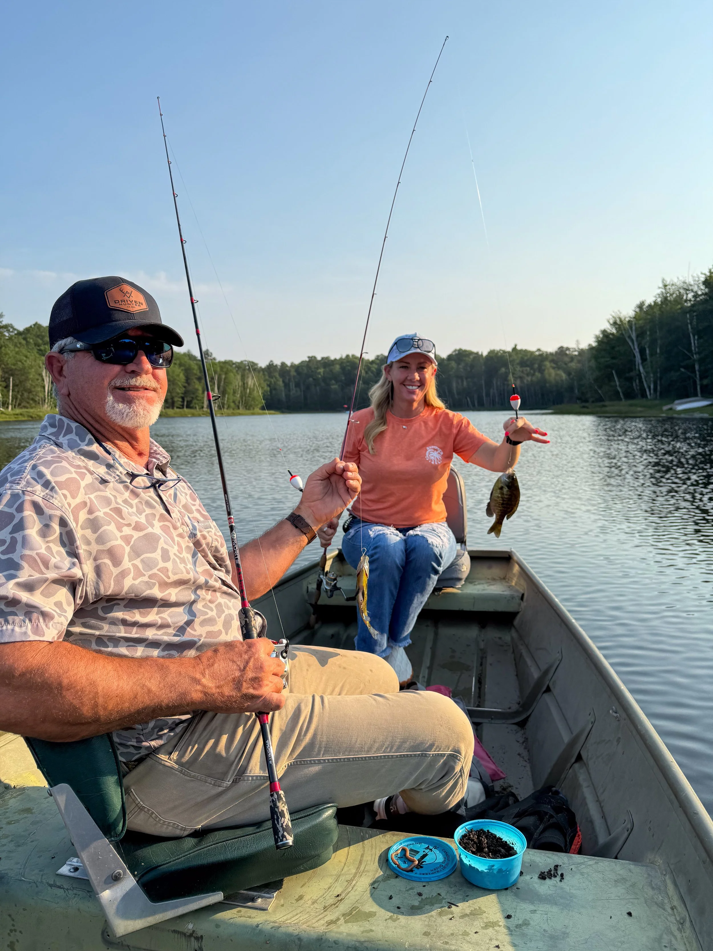 An older man and a woman sit in a small boat on a calm river, holding fishing rods with caught fish, surrounded by trees and a clear blue sky.
