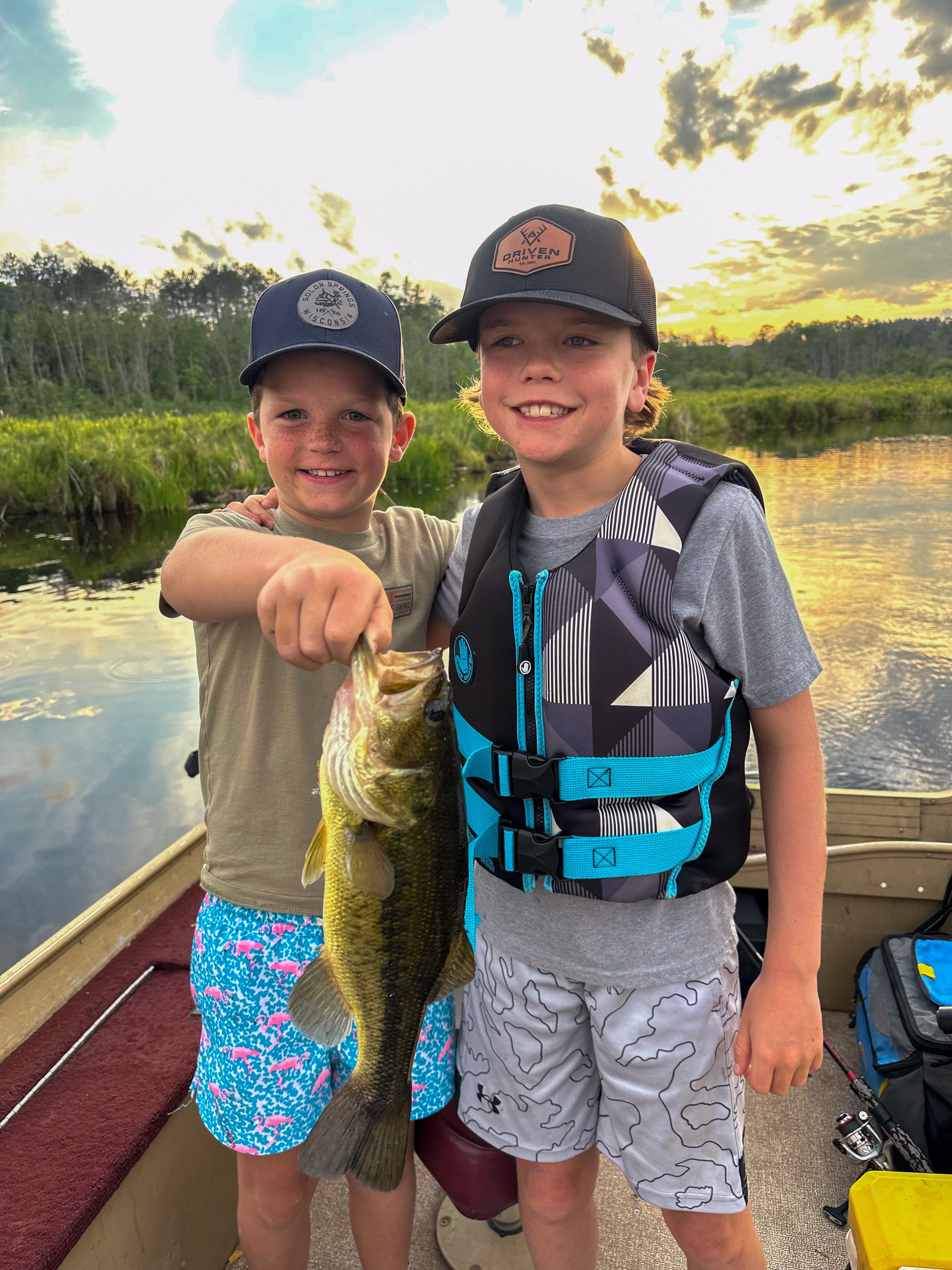 Two young boys standing on a boat holding a large fish they caught during sunset on a river, with trees and clouds in the background.