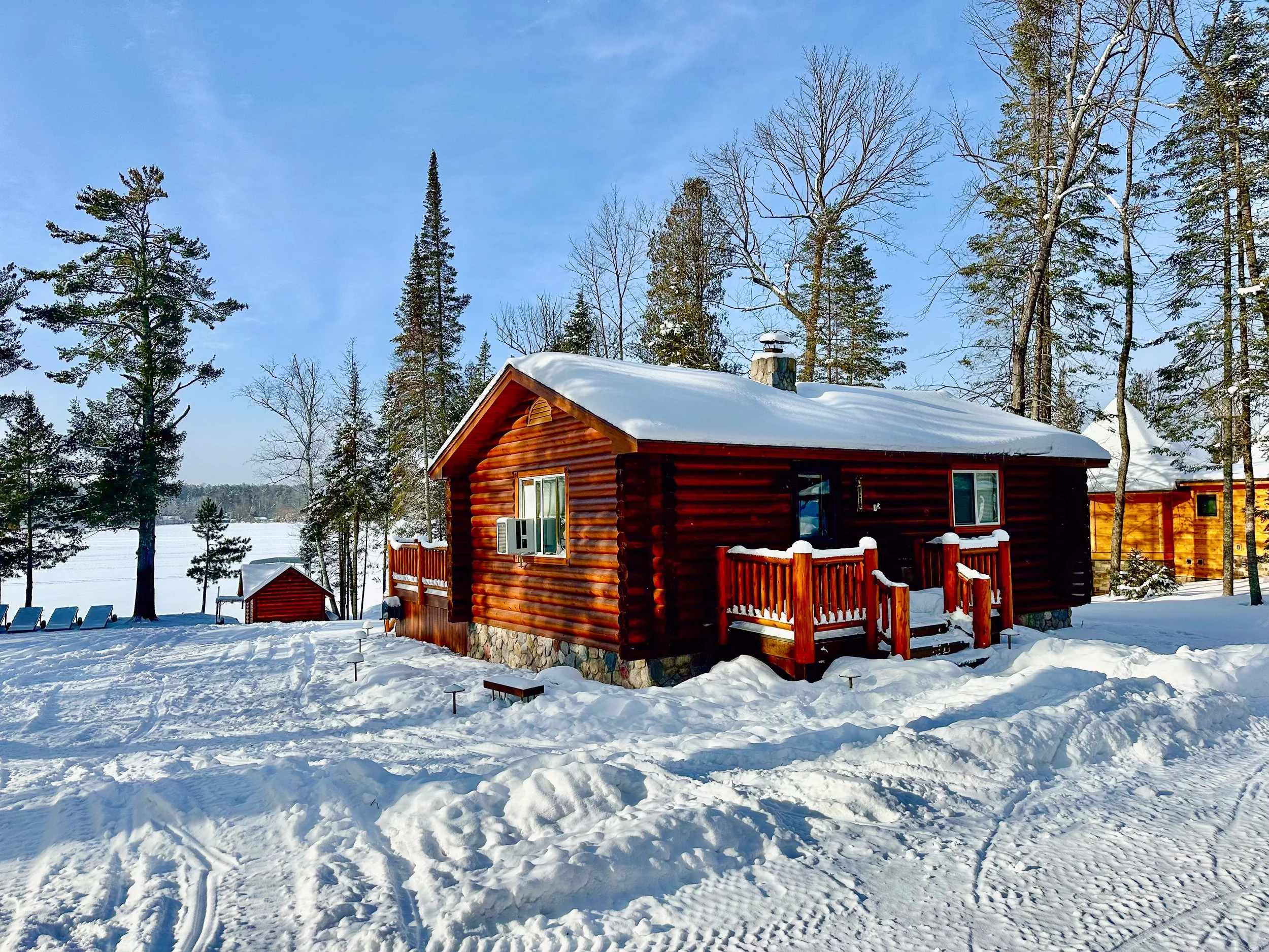 A snow-covered log cabin with a small porch in a winter landscape surrounded by tall trees and a frozen lake in the background.