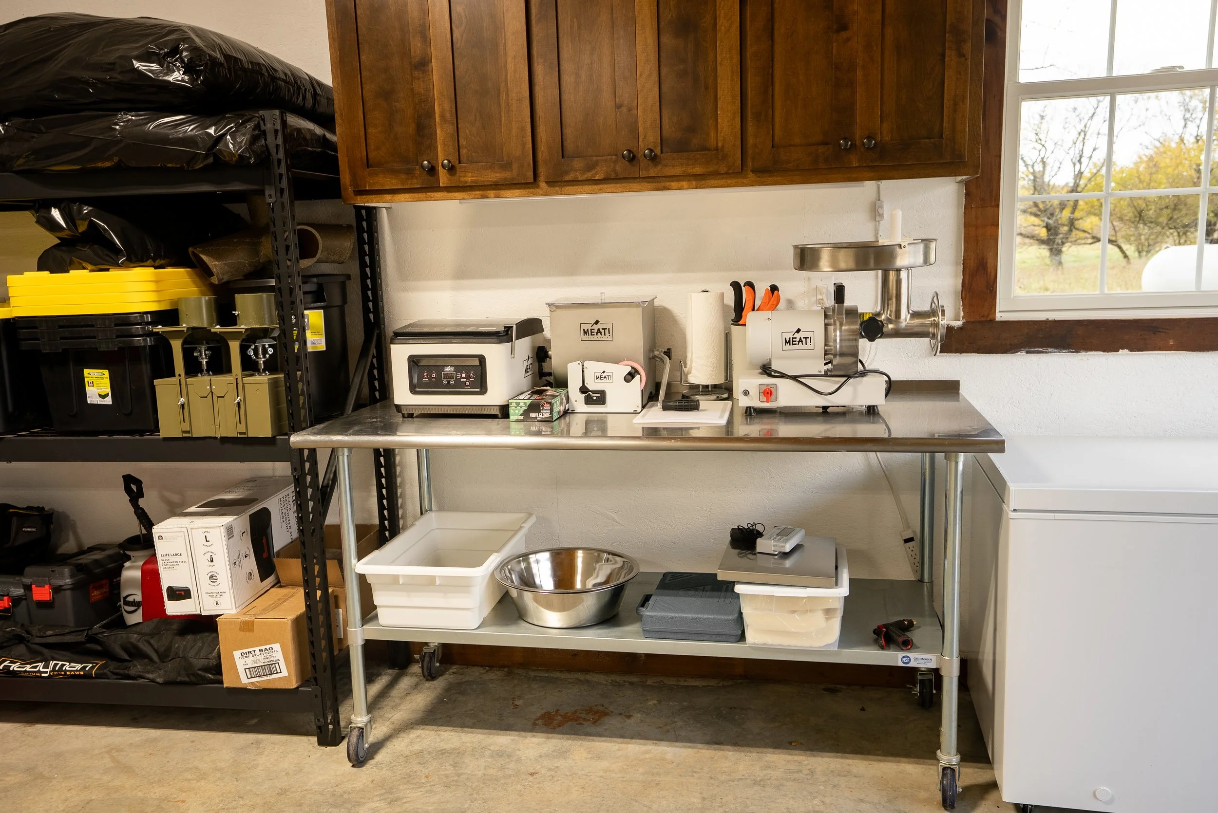 A stainless steel bench with a meat grinder, packaging equipment, and other kitchen appliances, with shelving on the left holding storage containers and equipment, and a window in the background revealing trees outside.