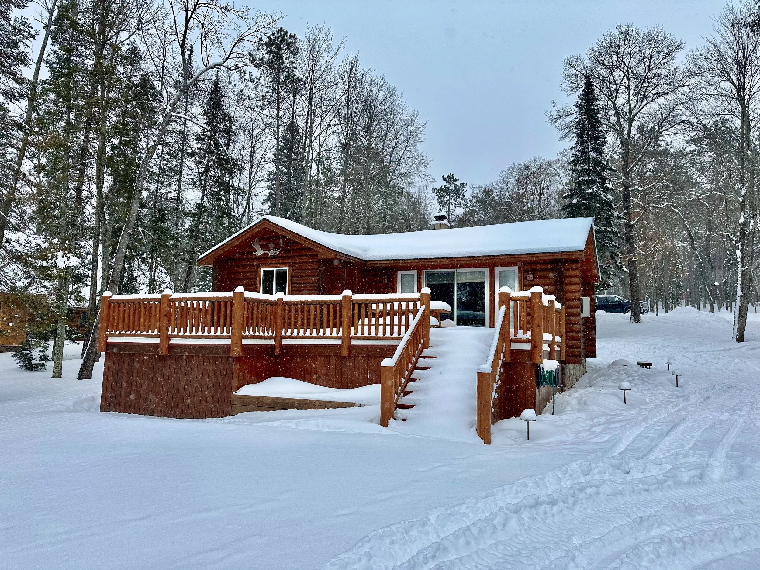 A wooden cabin with a large deck and stairs, surrounded by snow-covered ground and tall trees in a winter landscape.
