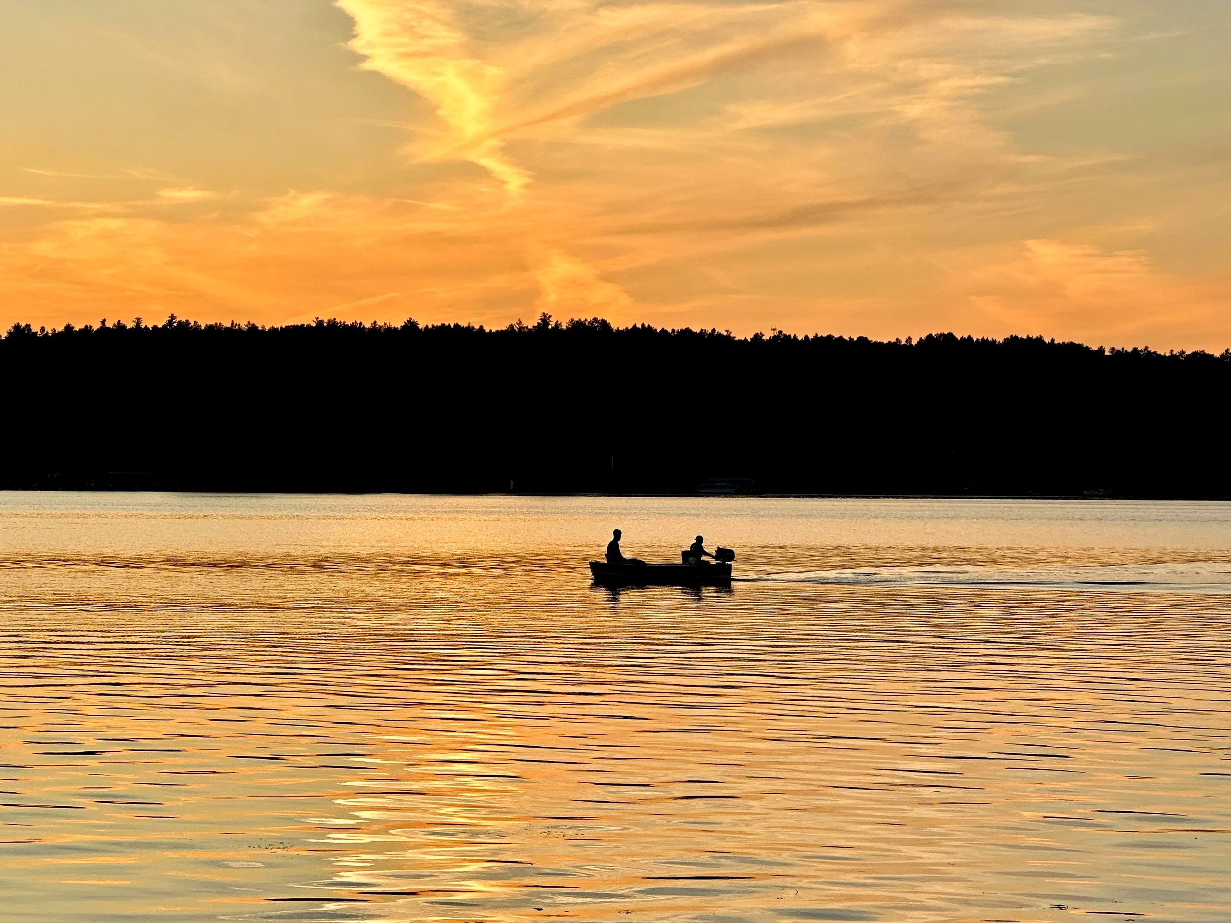 Silhouetted boat with two people on a calm lake during sunset, with a tree-covered shoreline and colorful sky.