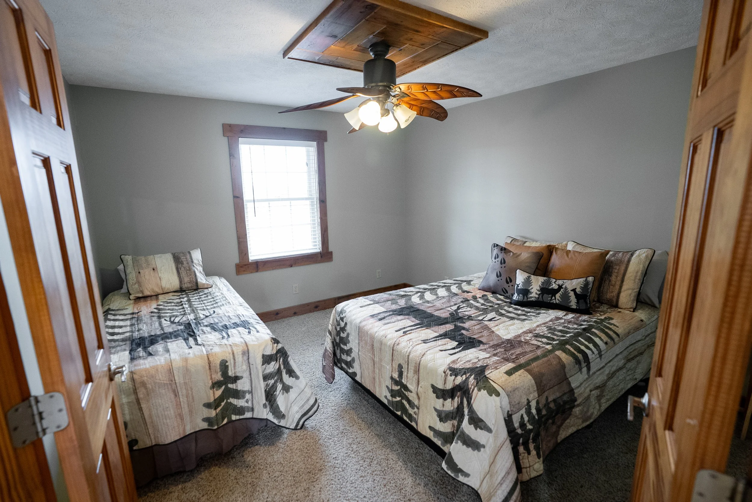 A bedroom with two beds, one smaller and one larger, both covered with rustic, nature-themed bedspreads featuring trees and animals. The room has light gray walls, a window with wooden trim, and a ceiling fan with wooden blades and a light fixture. T