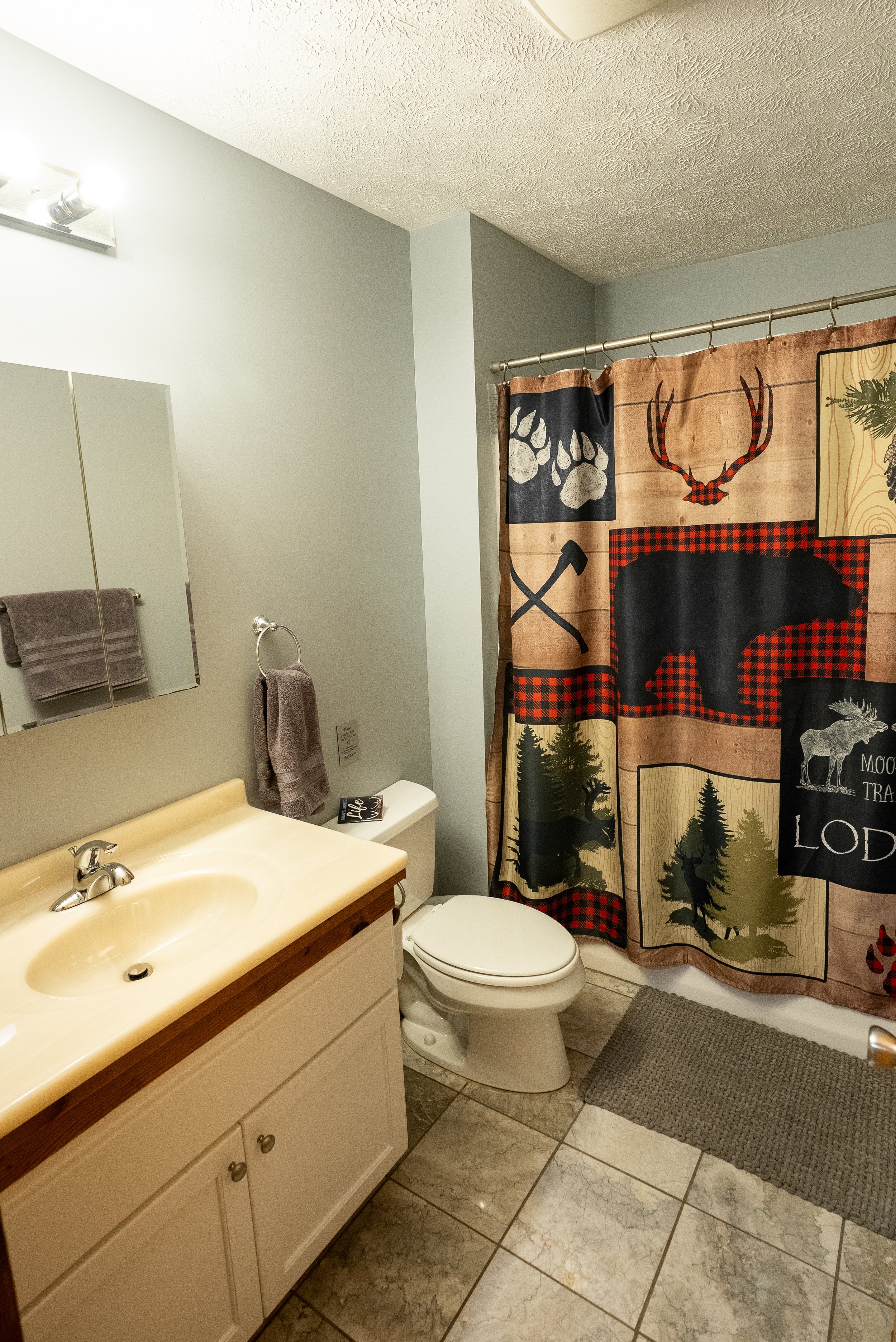 A bathroom with a vanity, mirror, towel, toilet, and a shower with a fabric curtain featuring a woodland design with a bear, moose, trees, and axes.