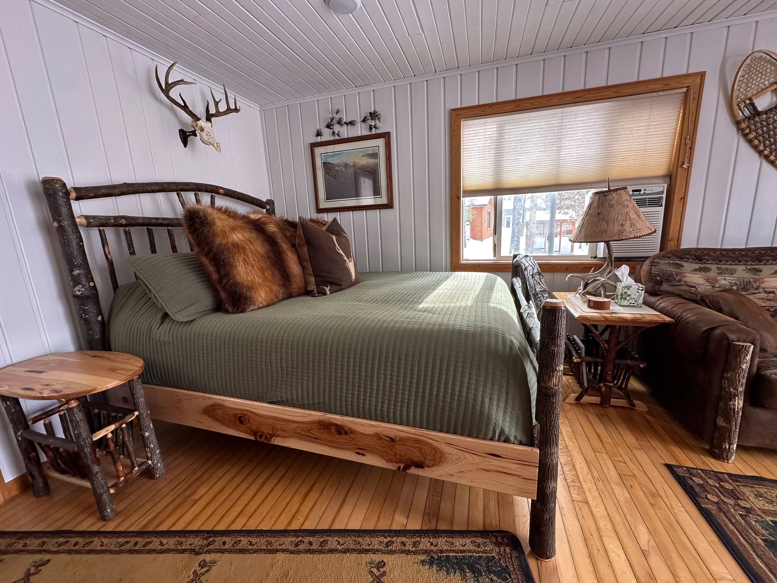 A rustic bedroom with a wooden log bed frame, green bedding, and decorative pillows. There is a small wooden side table and a window with a beige shade. The walls are white beadboard, with a mounted deer skull with antlers, a picture frame, and other