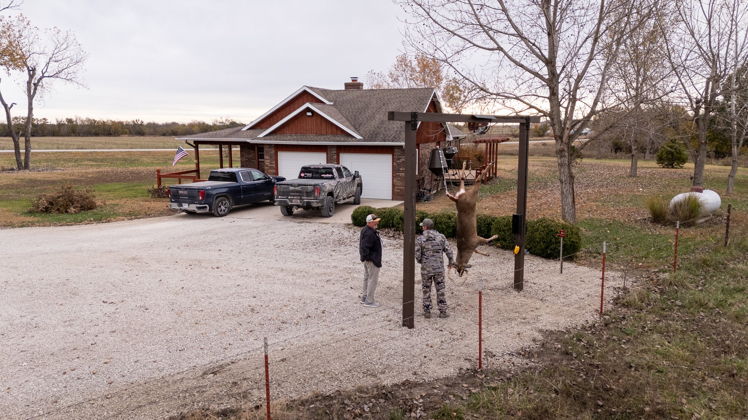 A rural property with a gravel driveway, two parked trucks, and a brick house with a garage. Two people are standing near a mounted animal decoy, a hanging pig carcass, in front of a wooded area. The scene appears to be during late fall, with some tr