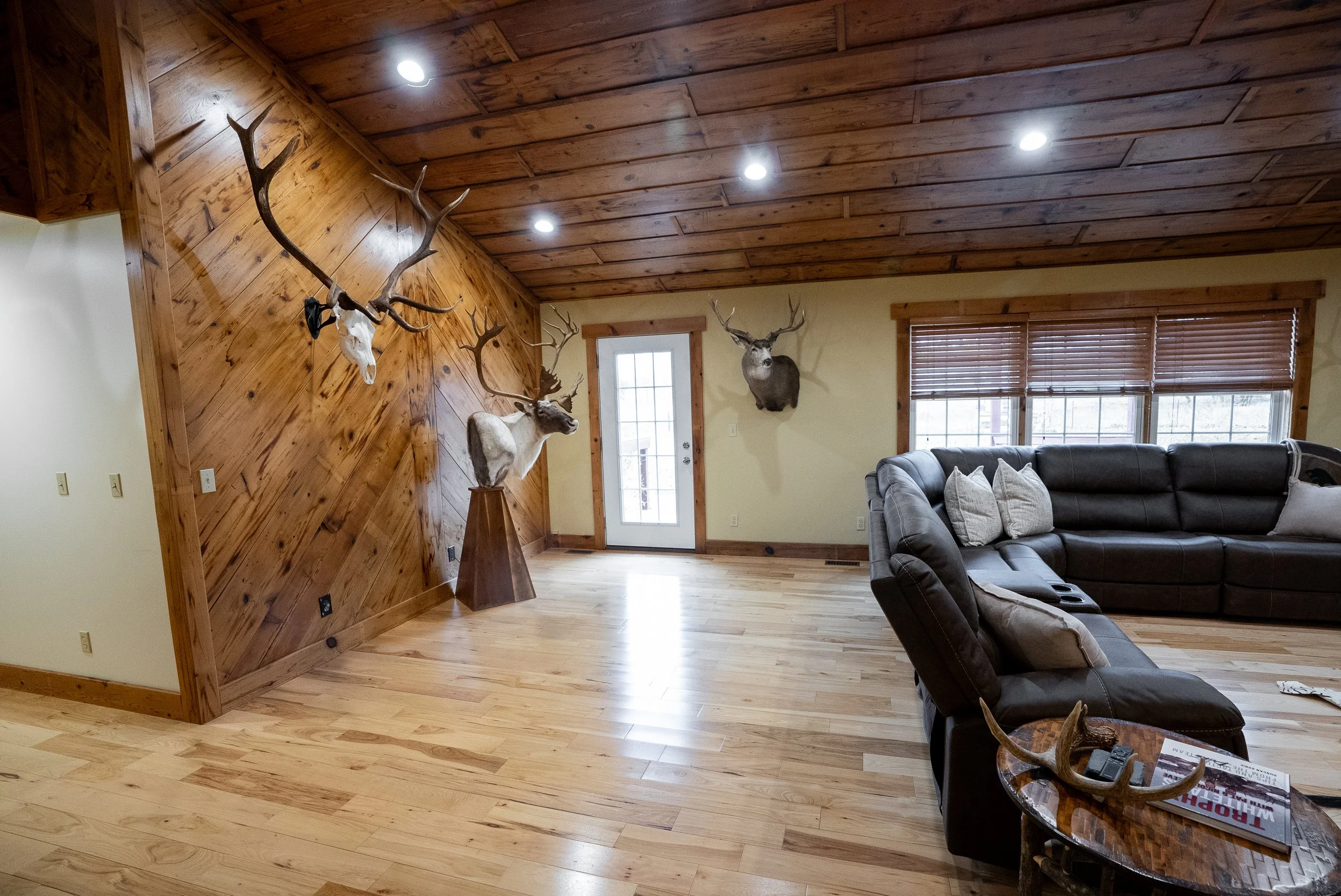 Living room with wooden accents, ceiling, and floor; animal head mounts on the wall including a deer with large antlers; black leather sectional sofa with pillows; small table with antler decor and magazines; glass door and window with blinds.