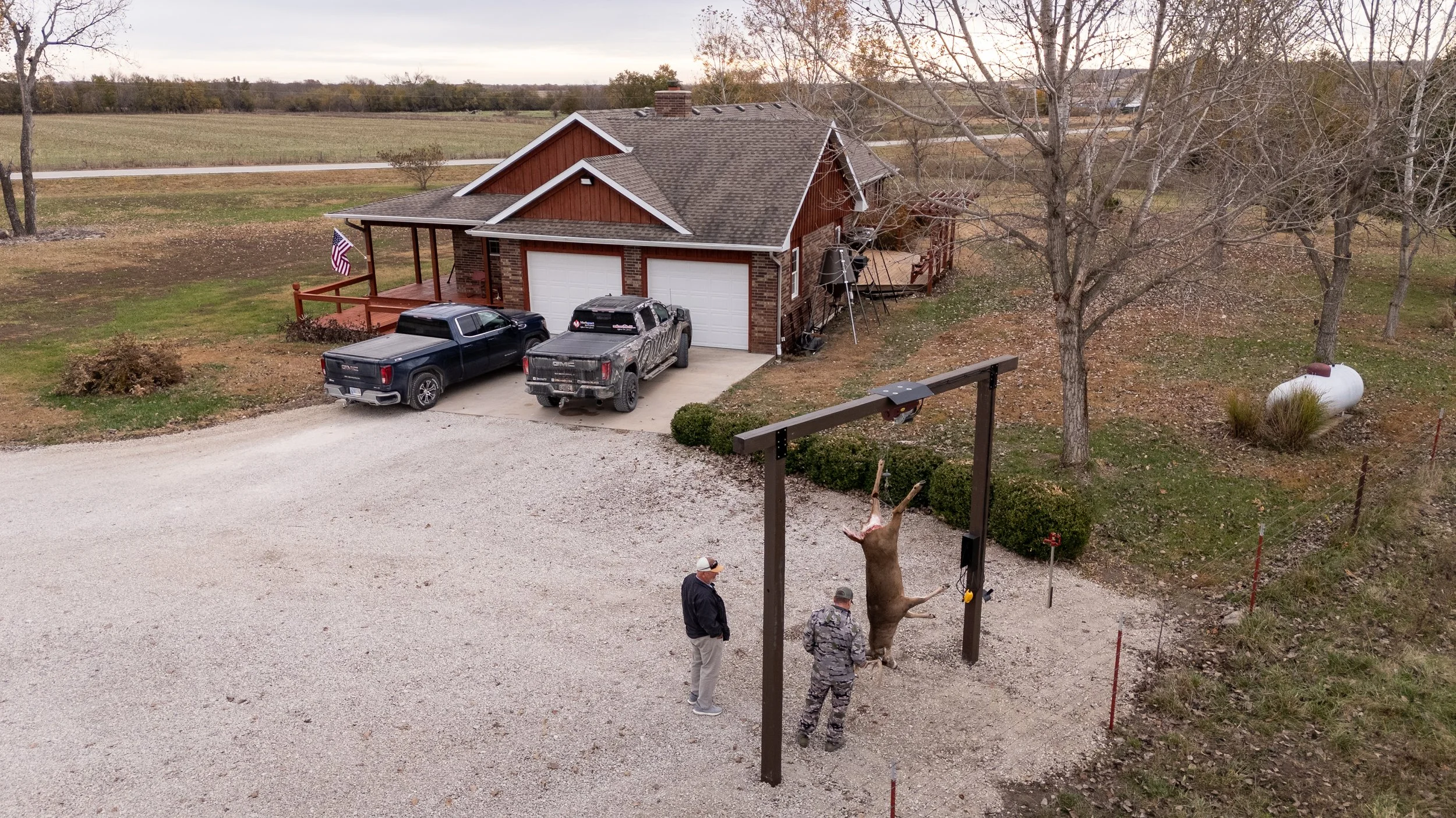 Two men stand under a wooden gallows with a hanging deer. A house with a brick exterior and three garage doors is in the background, along with three parked trucks and leafless trees.