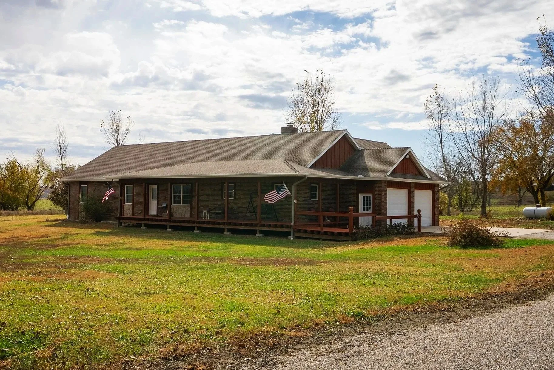 A single-story brick house with a front porch, American flags, and two garage doors, set on a grassy yard with trees and a cloudy sky in the background.