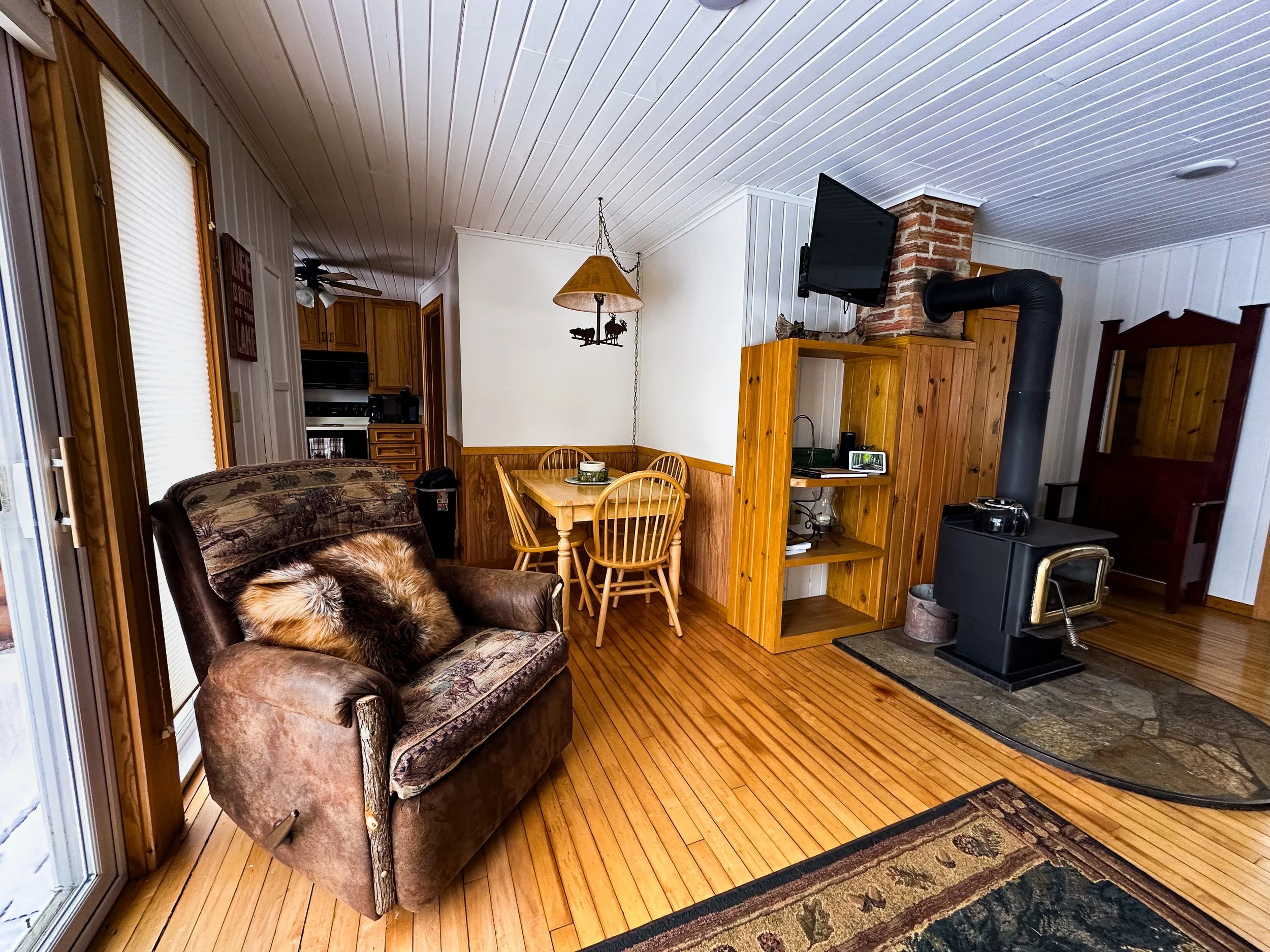 Living room with a brown armchair, a wooden dining table with four chairs, a wood stove, a flat-screen TV mounted on the wall, and wood paneling on the walls and ceiling.
