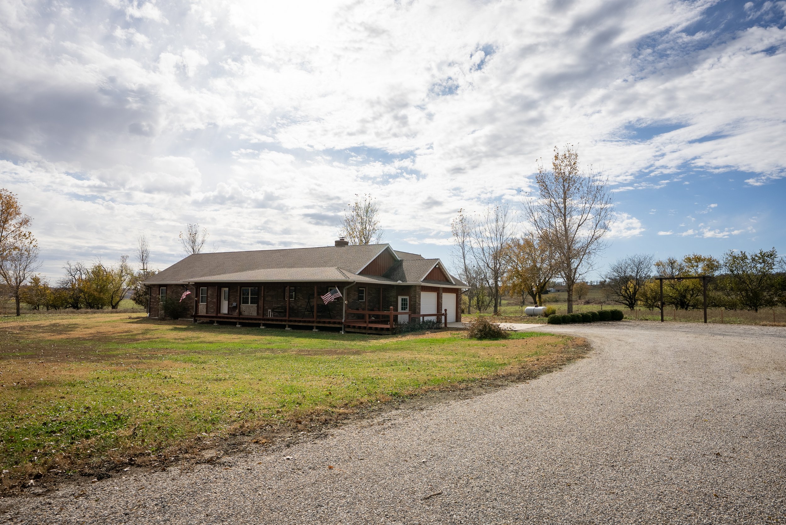 A house with a front porch and American flags, situated on a large grassy lot with trees and a gravel driveway under a partly cloudy sky.