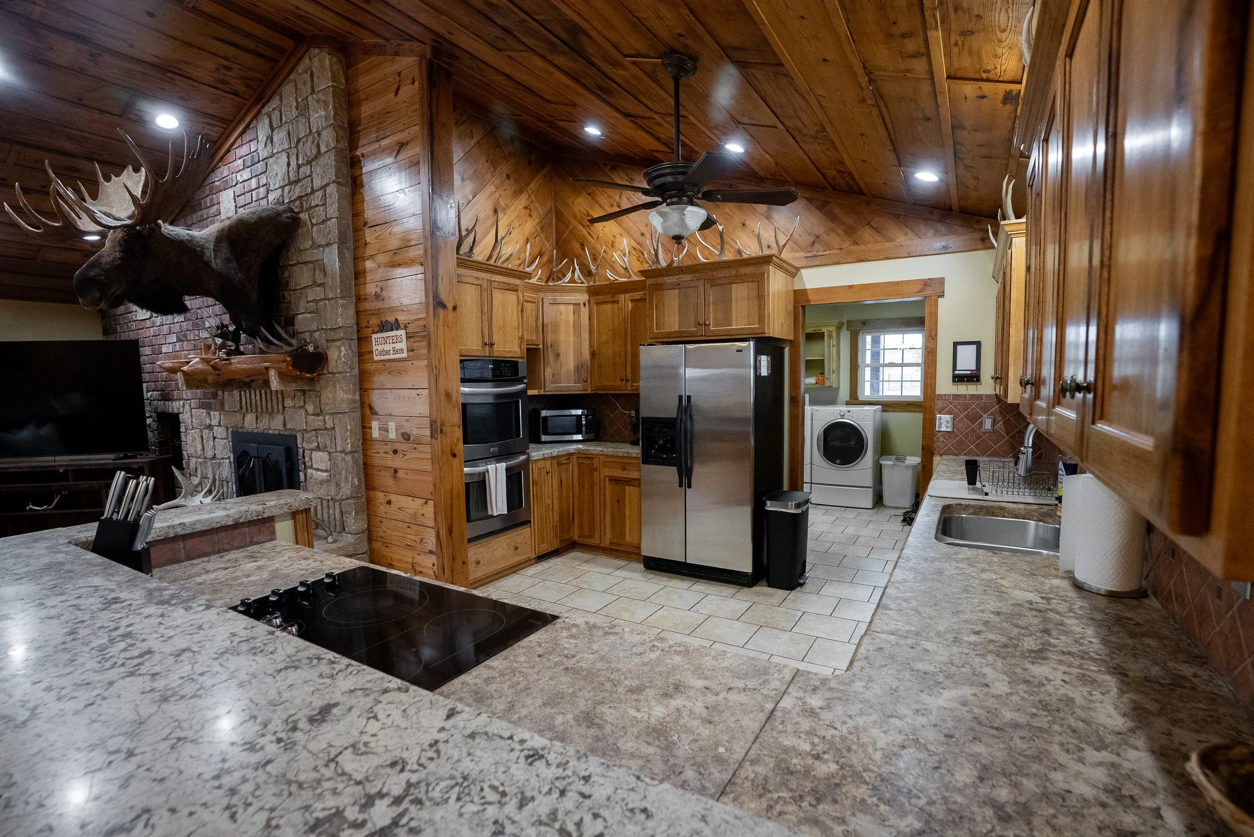 A rustic kitchen with wooden cabinets, a stainless steel refrigerator, and a stone fireplace with mounted moose and deer antlers. The kitchen has a granite countertop, a ceiling fan, and a washer and dryer in a nearby laundry area.