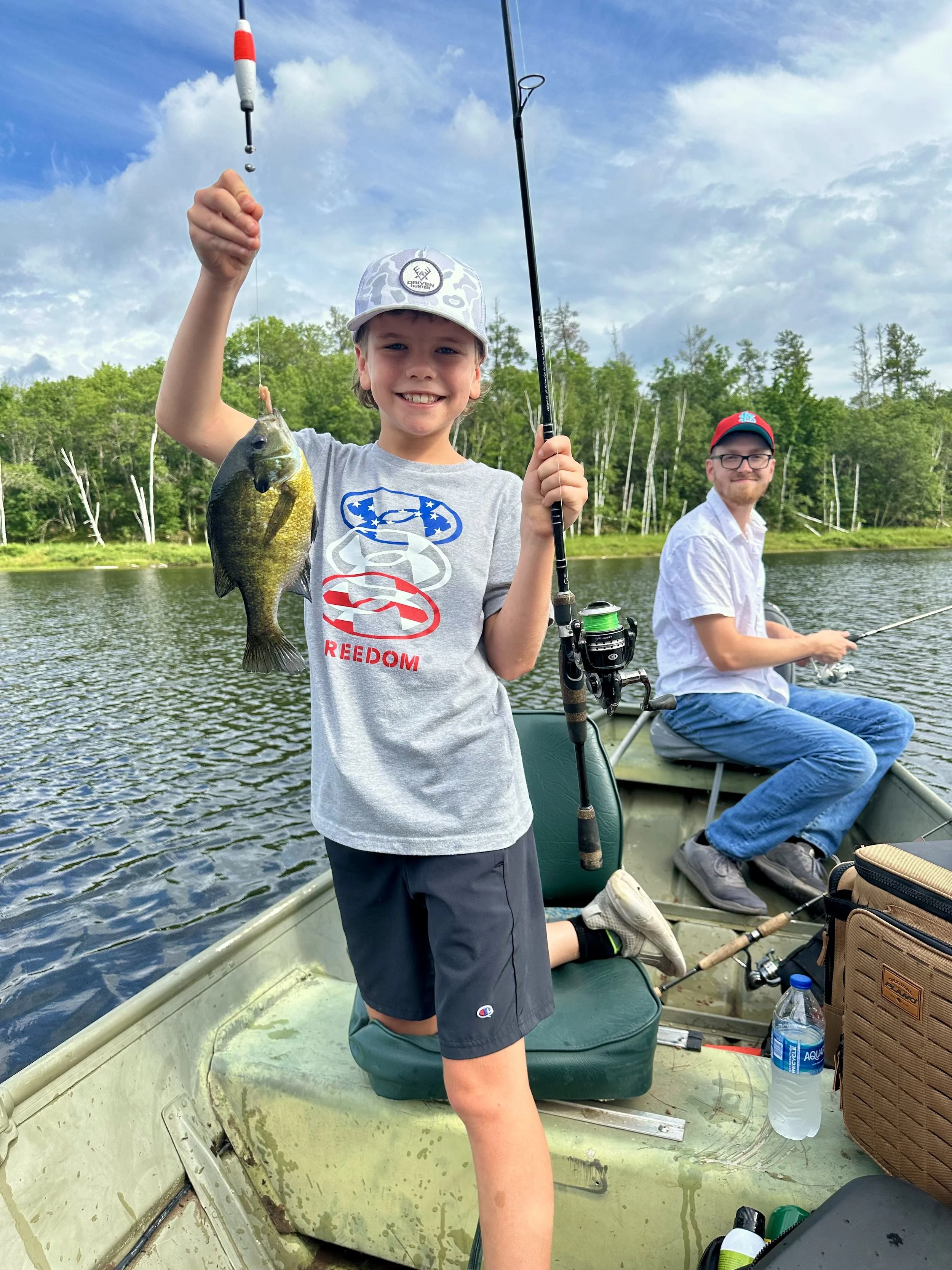 A young boy holding a fishing rod with a fish he caught, smiling, on a boat with a man fishing in the background, set on a river with trees and a partly cloudy sky.