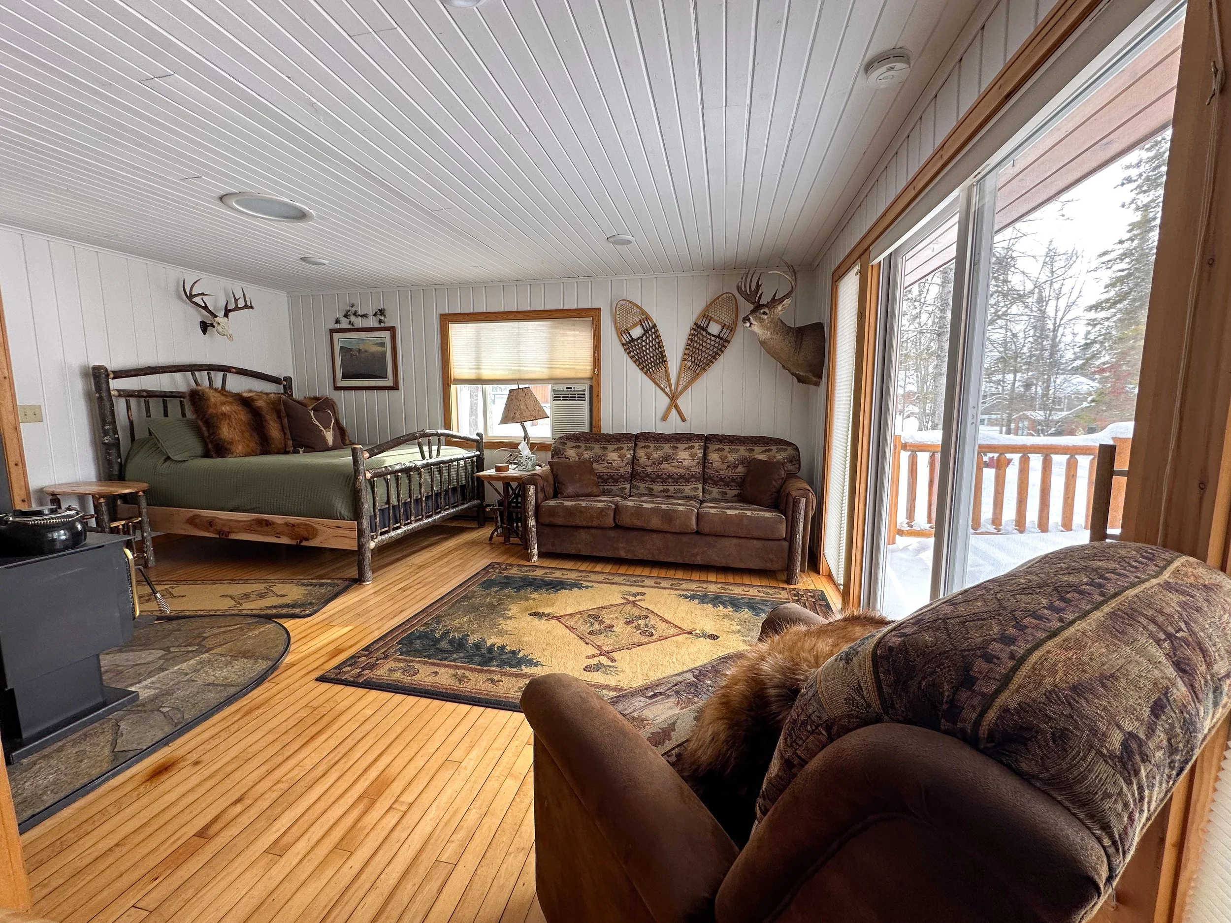 Cozy cabin living room with wood-paneled ceiling and walls, featuring a bed, a brown sofa, and a large sliding glass door leading to a snowy outdoor deck. Decor includes mounted deer heads, snowshoes, a patchwork area rug, and hunting trophies.