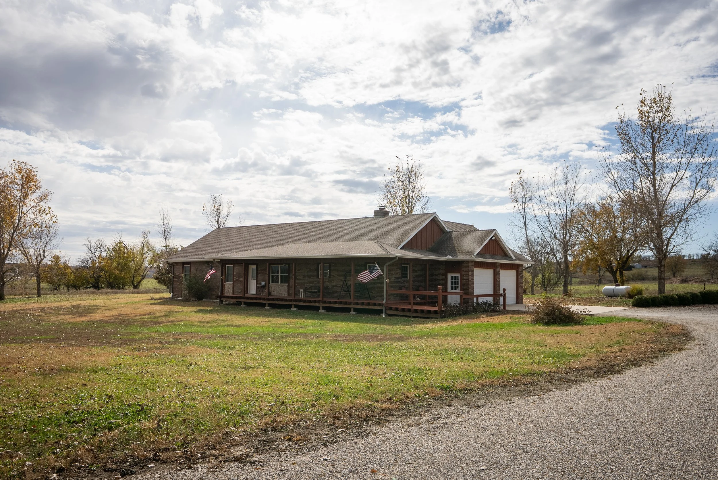 A single-story house with a brick and wood exterior, a front porch, and two attached garages. The house is surrounded by trees with some fall foliage, and a gravel driveway curves toward the garages. The sky is partly cloudy with patches of blue.
