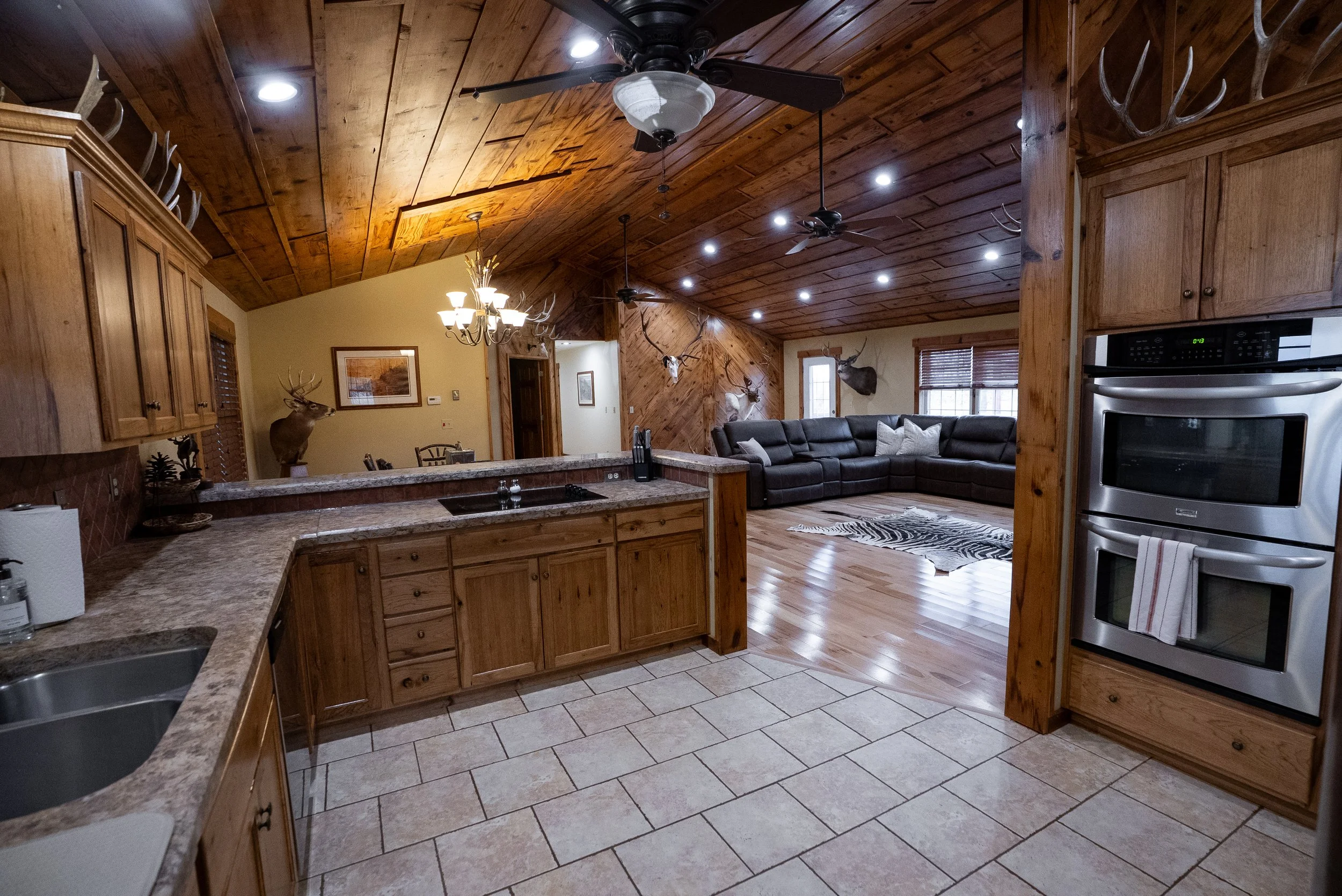 Open-concept kitchen and living room with wooden ceiling and walls, mounted deer antlers, mounted animal heads, black sectional sofa with pillows, zebra print rug, stainless steel double oven, tiled kitchen floor, and hardwood living room floor.