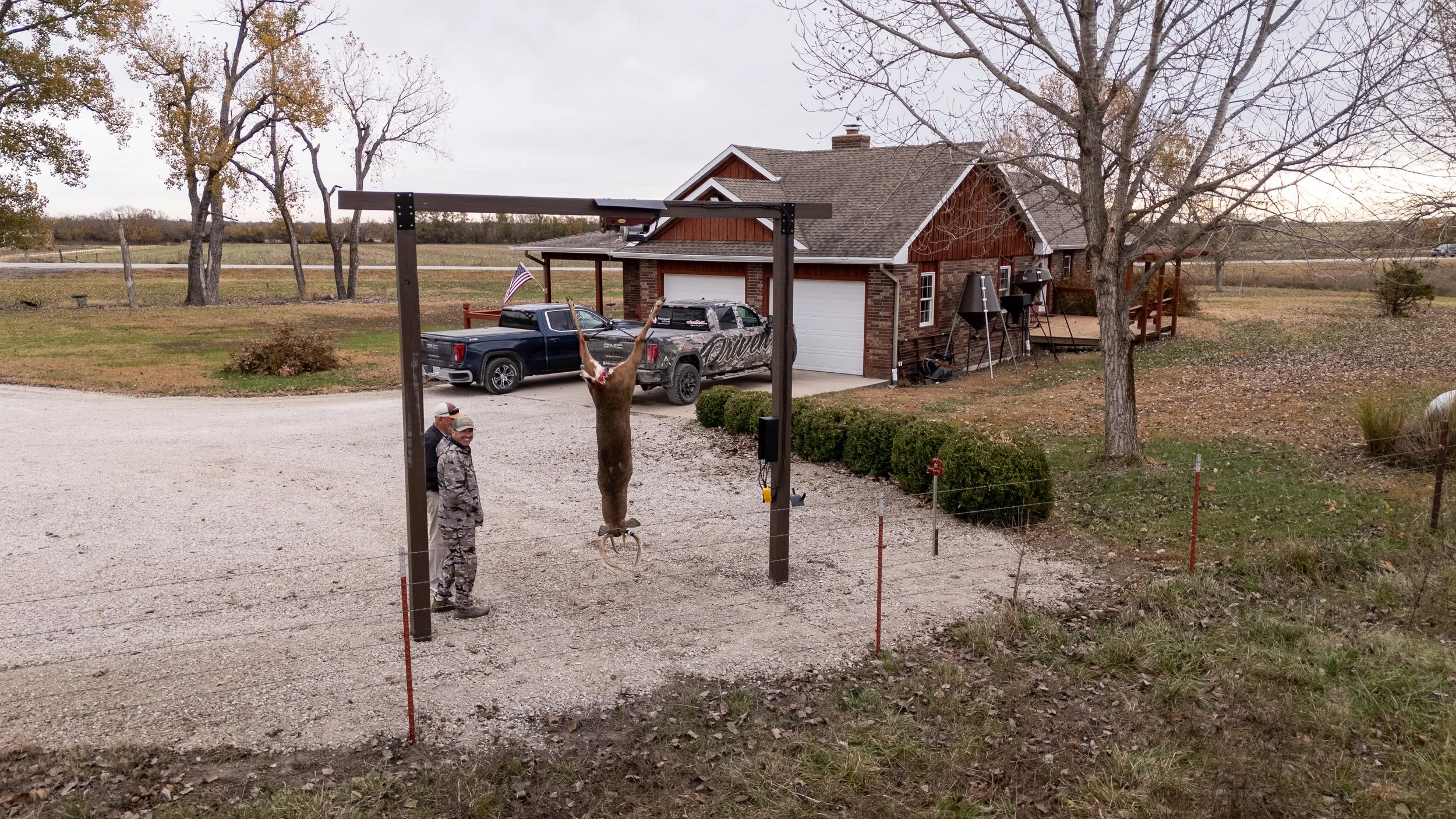 A person hanging a large buck carcass upside down from an outdoor metal frame, with two men observing. There are two trucks parked in the driveway near a house, with trees and an open field in the background.