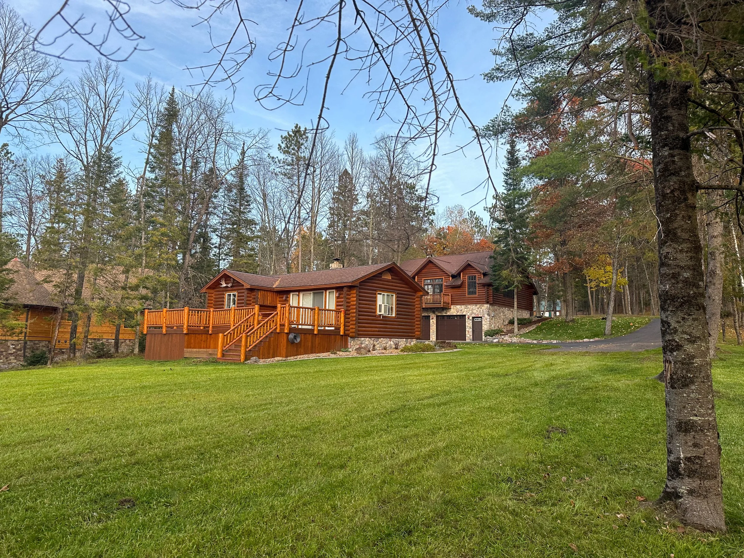 A wooden house with a deck and stairs, surrounded by a green lawn and tall trees, with a paved driveway leading to the house.