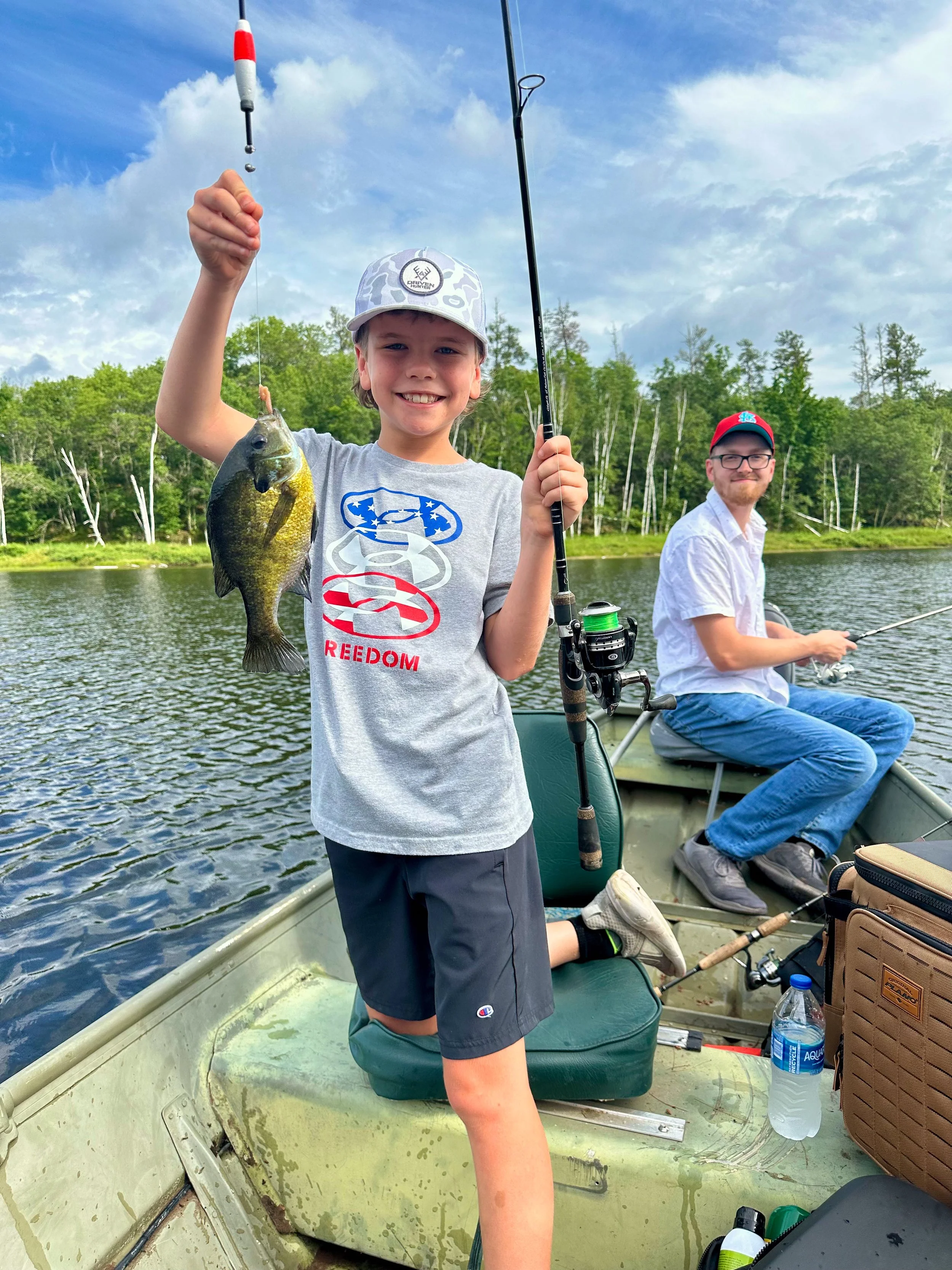 A young boy smiling and holding a fishing rod with a fish he caught on a boat in a lake, with a man sitting in the background fishing and a green forested shoreline behind them.