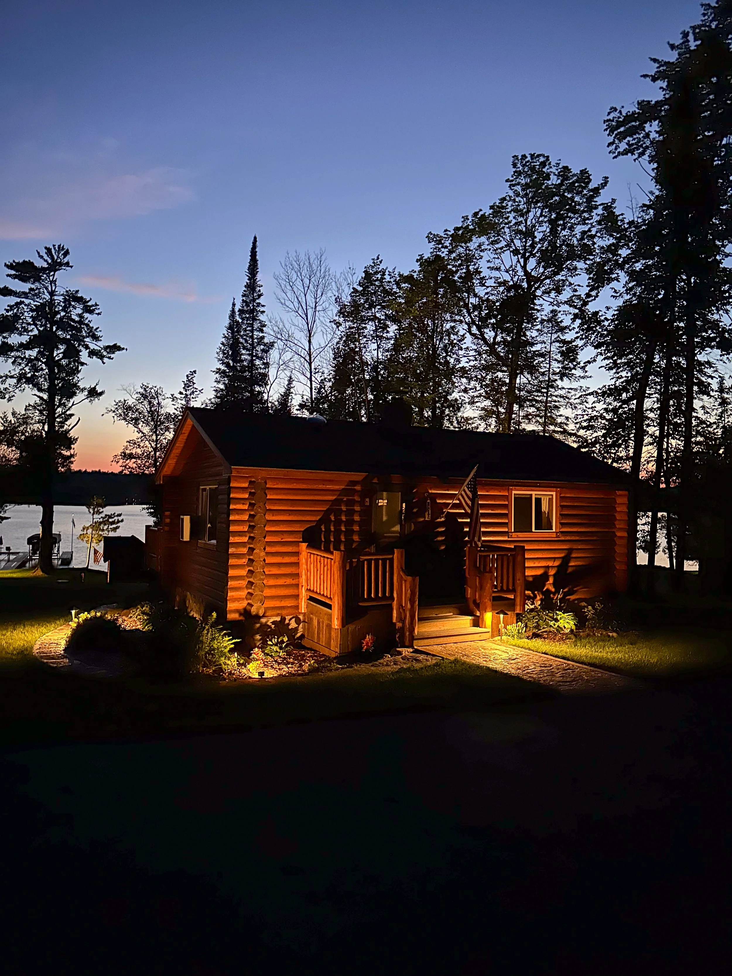 A wooden cabin illuminated by outdoor lighting at dusk, surrounded by tall trees and near a body of water.