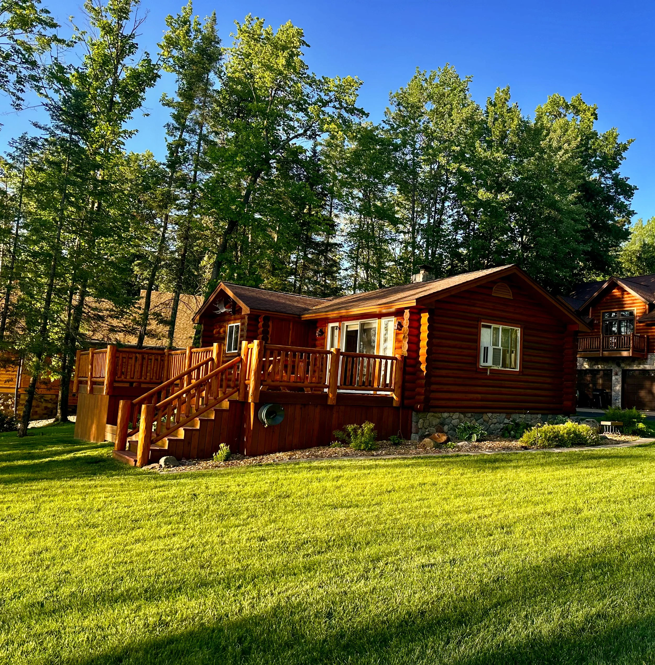 A wooden house with a deck, surrounded by lush green grass and tall trees under a bright blue sky.