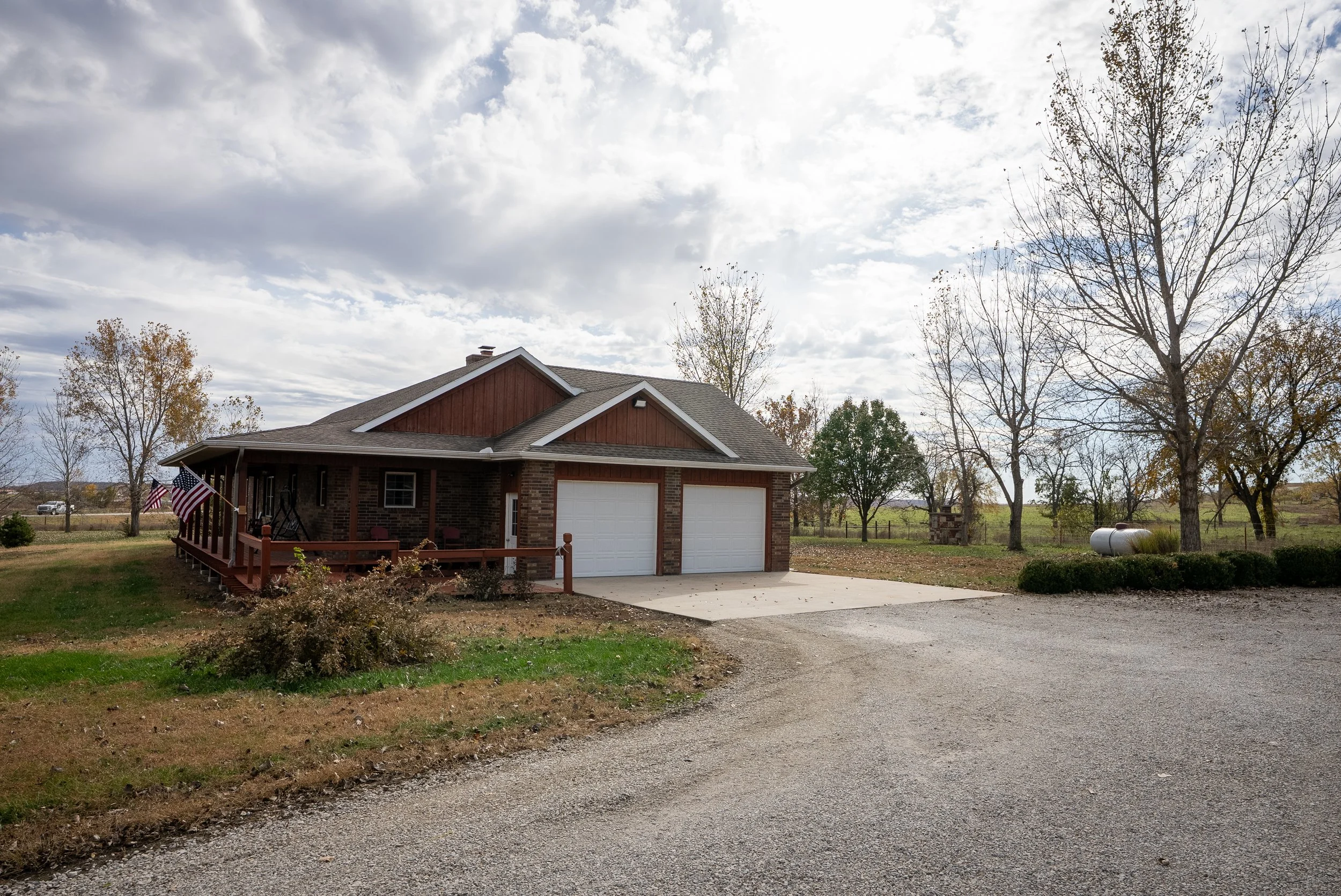 A single-story house with a brick facade, front porch, and two white garage doors, situated in a rural setting with leafless trees and a cloudy sky.
