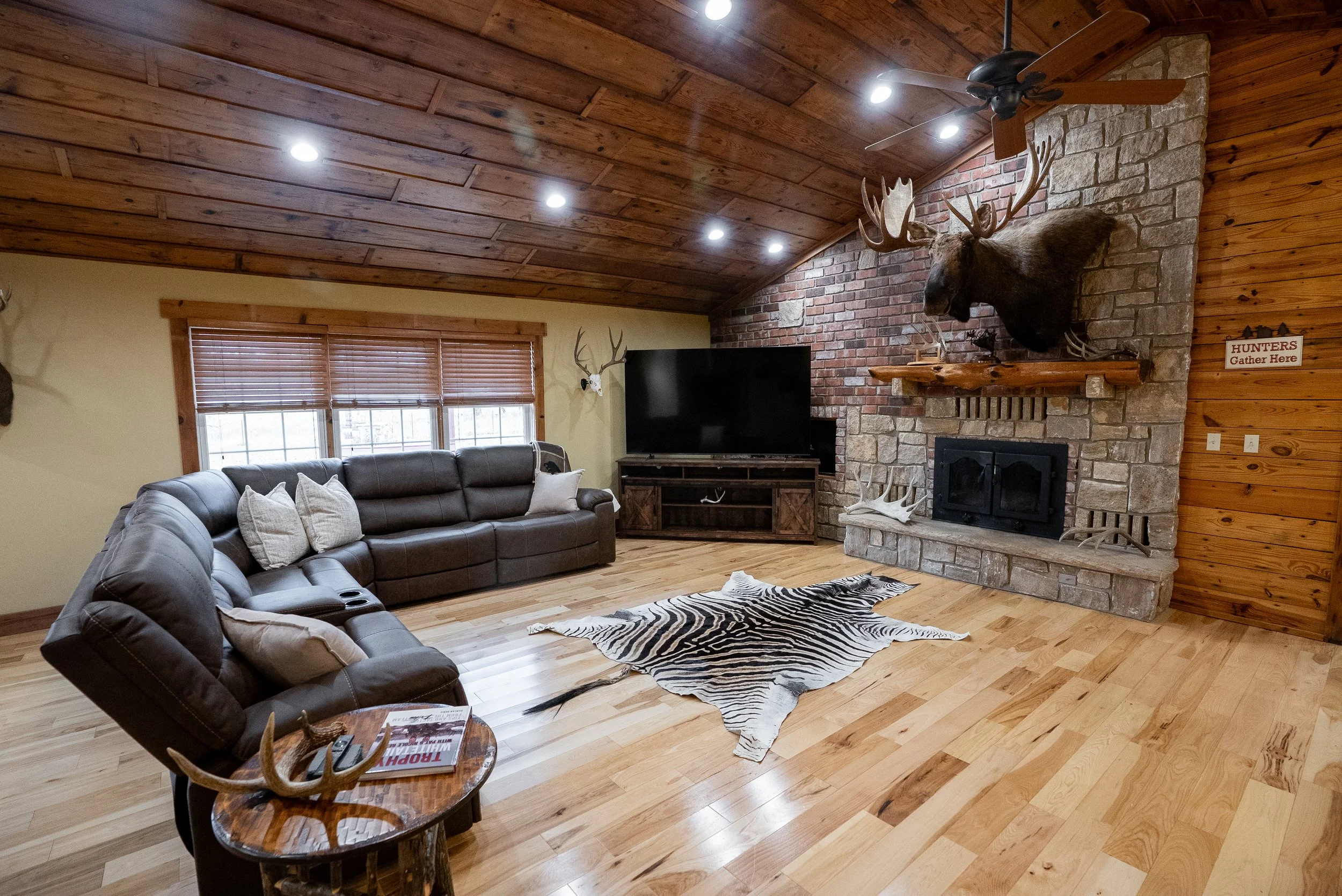Living room with wooden ceiling and floors, brick fireplace with mounted moose head, brown leather sectional sofa, zebra-patterned rug, window with wooden blinds, flat-screen TV on wooden stand, and antlers on side wall.