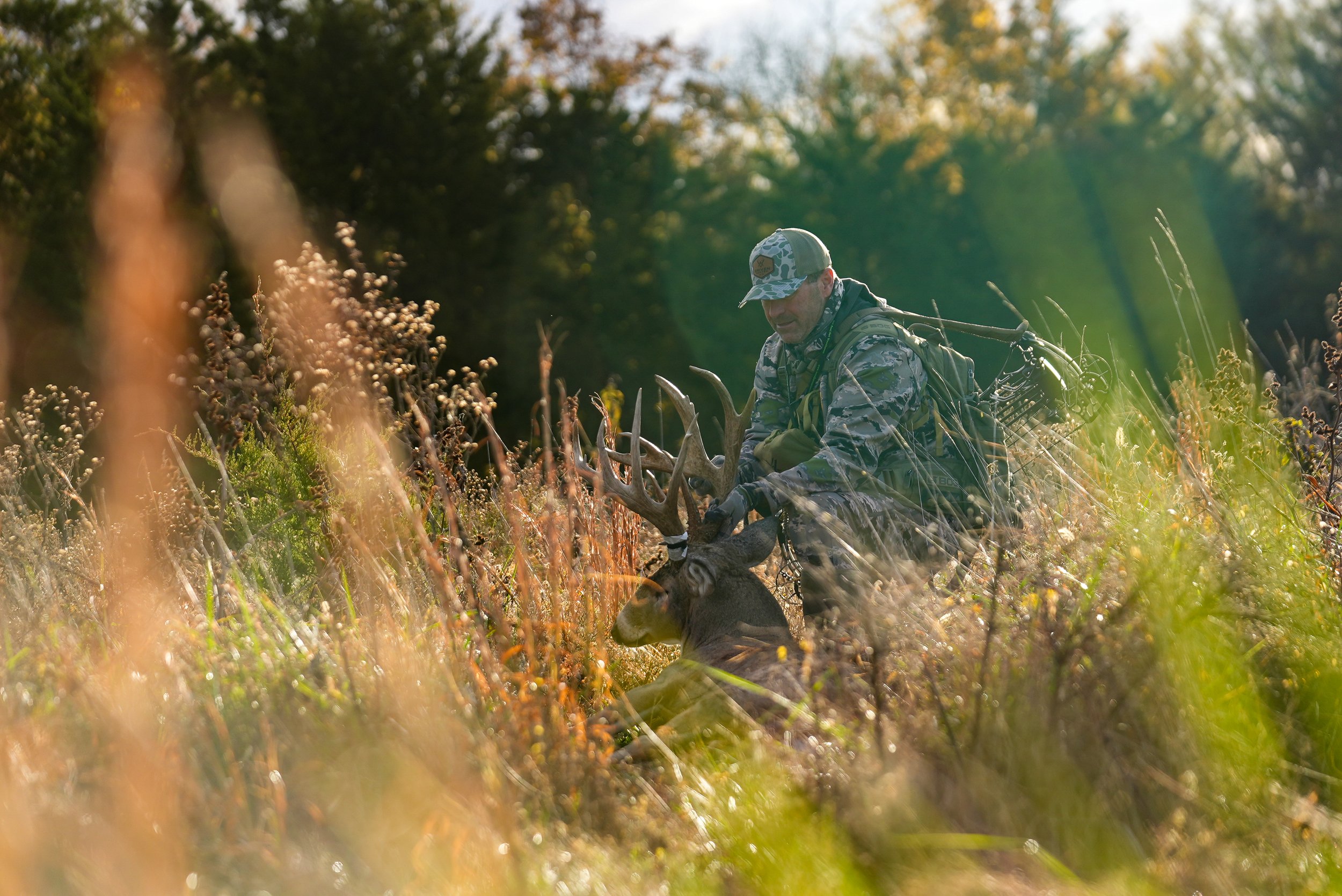 A man in camouflage hunting gear kneels in a field of tall grass and flowers, holding the antlers of a large buck with multiple points.