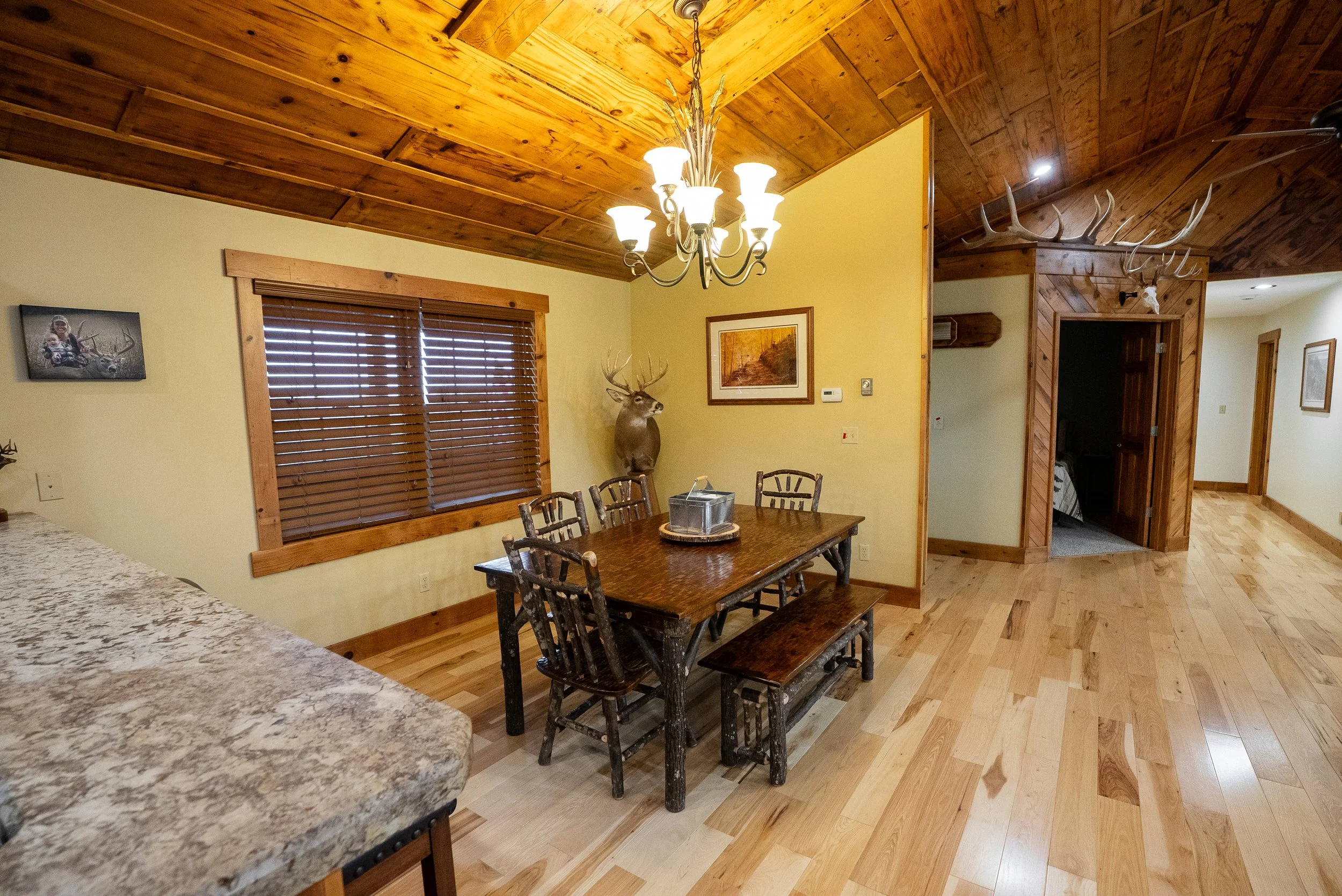 A rustic dining room with wooden ceilings, walls, and floors. There is a wooden dining table with chairs and a bench, a chandelier above, and a mounted deer head on the wall.