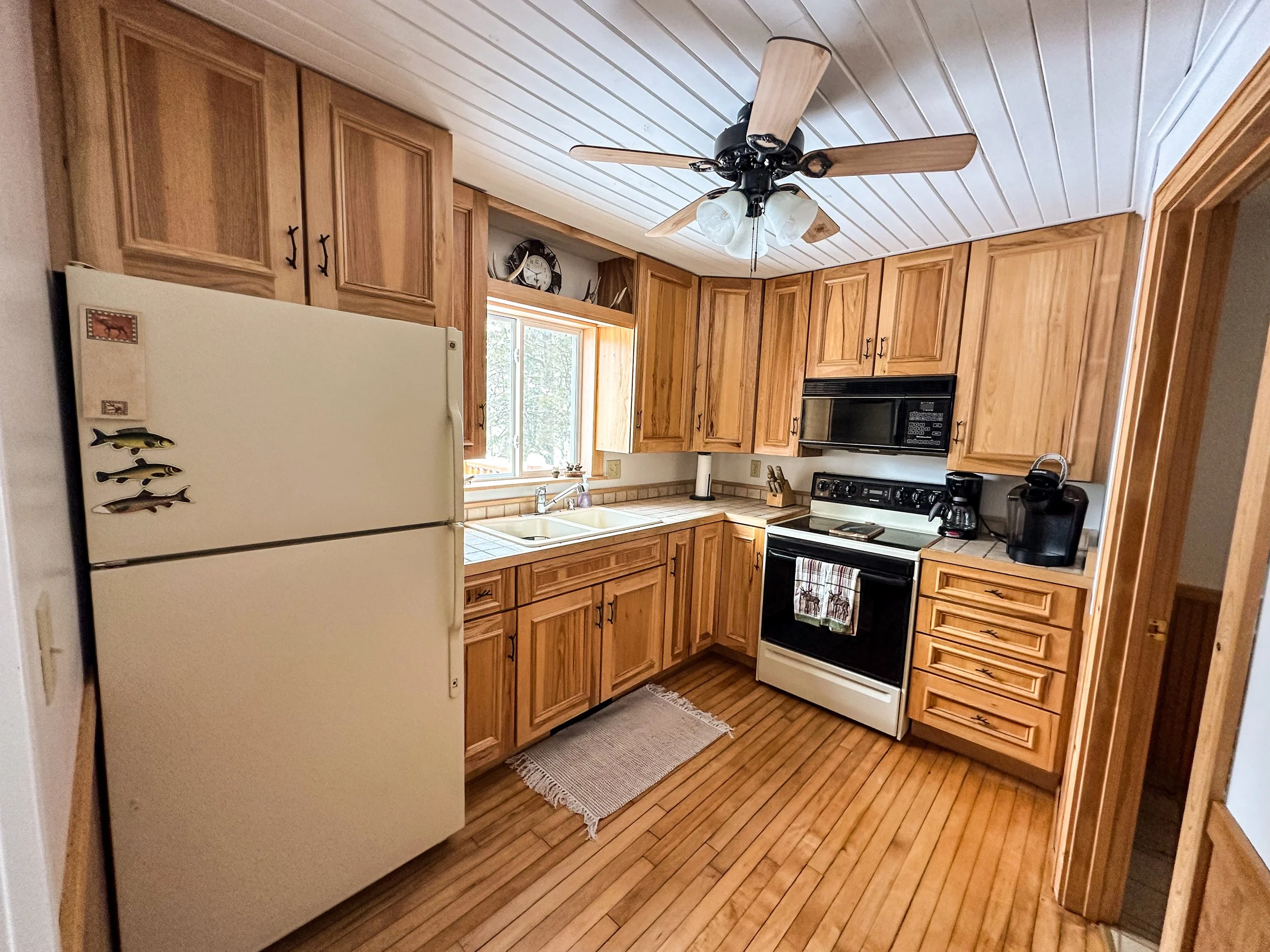 Kitchen with wooden cabinets, white refrigerator with fish magnets, black microwave, oven, coffee maker, and ceiling fan with lights, window above sink with natural light.