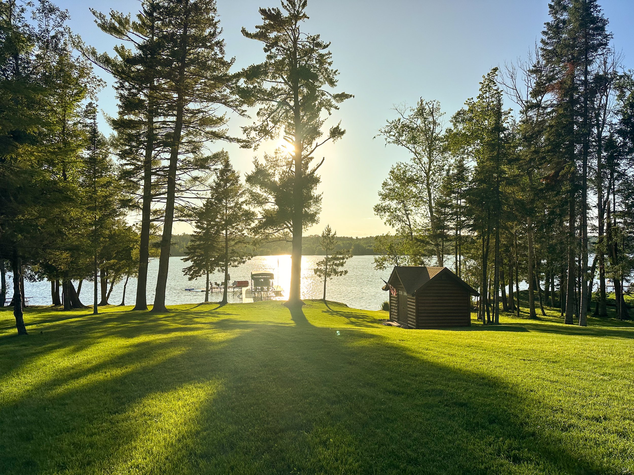 Sunset over a lake with trees and a small wooden shed in the foreground.