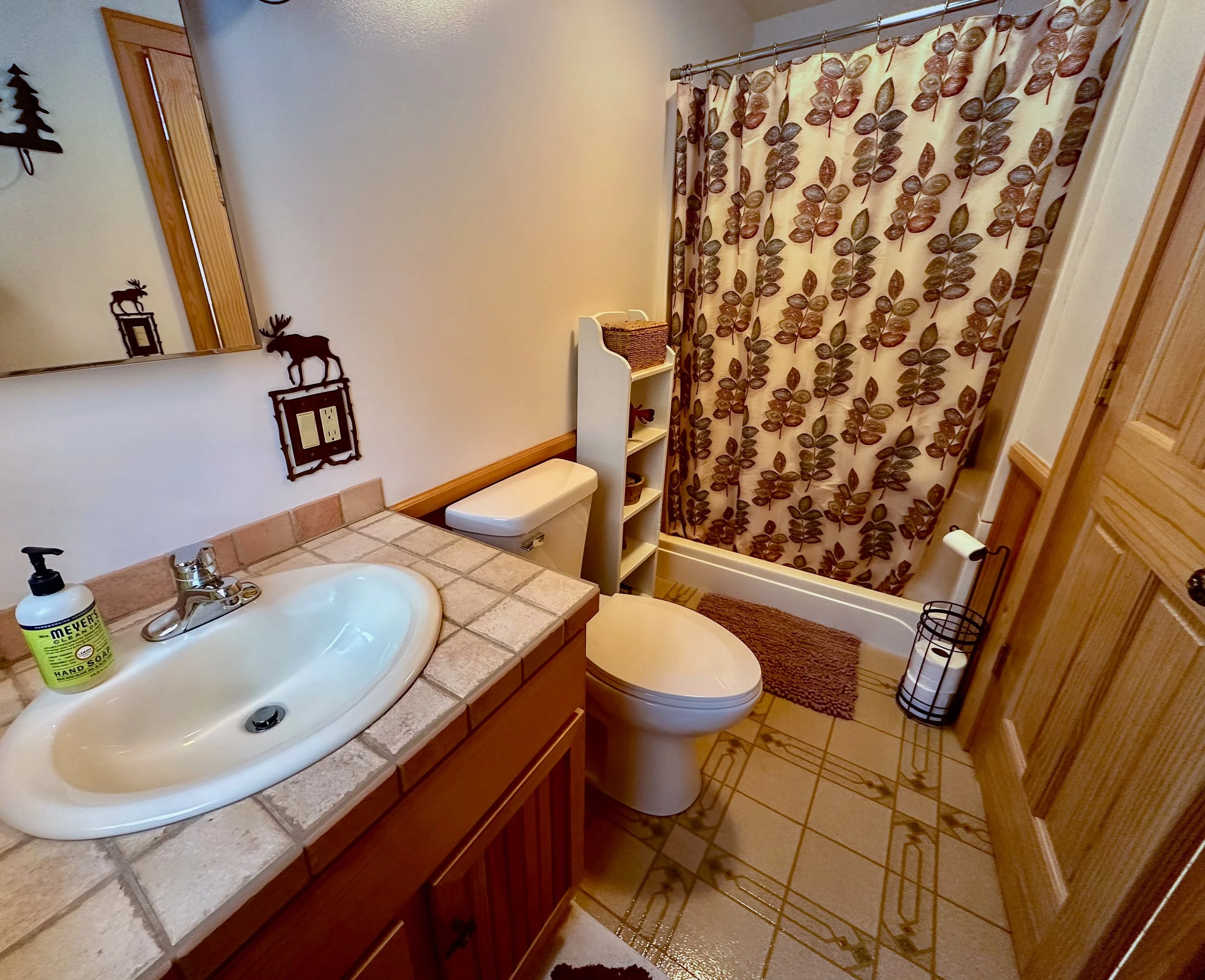 A small bathroom with a sink, toilet, and shower. The sink has a beige tile countertop with a soap dispenser. The shower has a curtain with brown leaves pattern. There is a mirror above the sink, a wooden cabinet on the right, and a white shelf with 