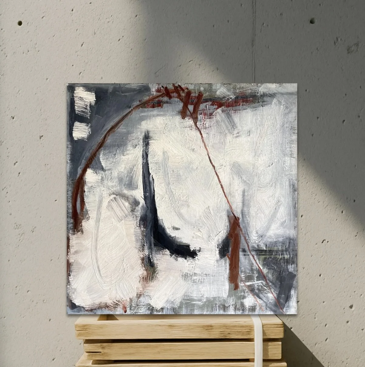 Abstract painting with white, black, gray, and brown brushstrokes on a canvas, placed on a wooden stool against a textured wall.