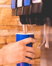 A hand holding a blue cup under a soda fountain dispensing drink