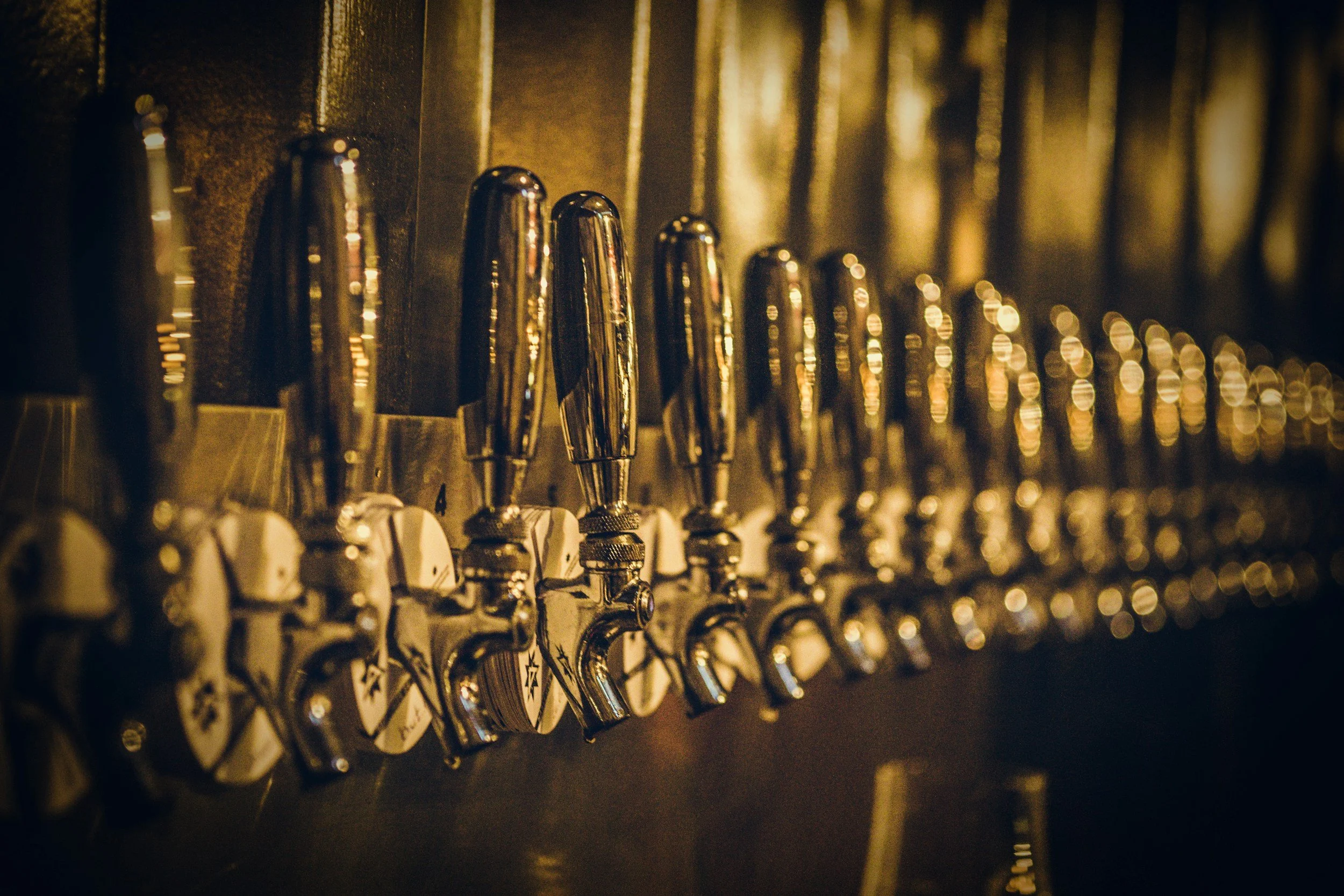 Multiple beer taps lined up on a dark wooden bar with warm lighting.