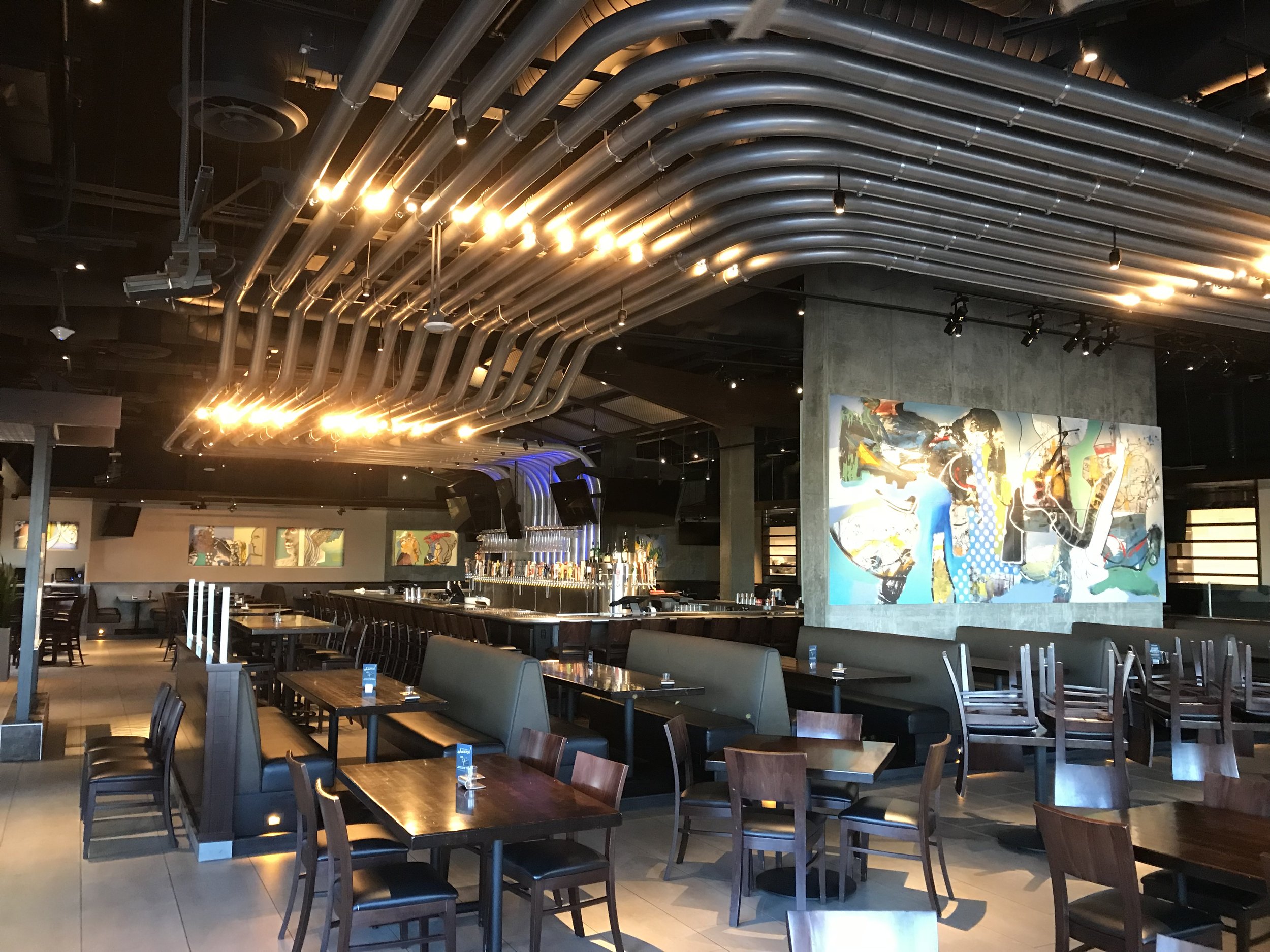 Interior of a modern restaurant or bar with wooden tables and chairs, artwork on the walls, and a bar area with bottles and taps, illuminated by warm and ambient lighting.