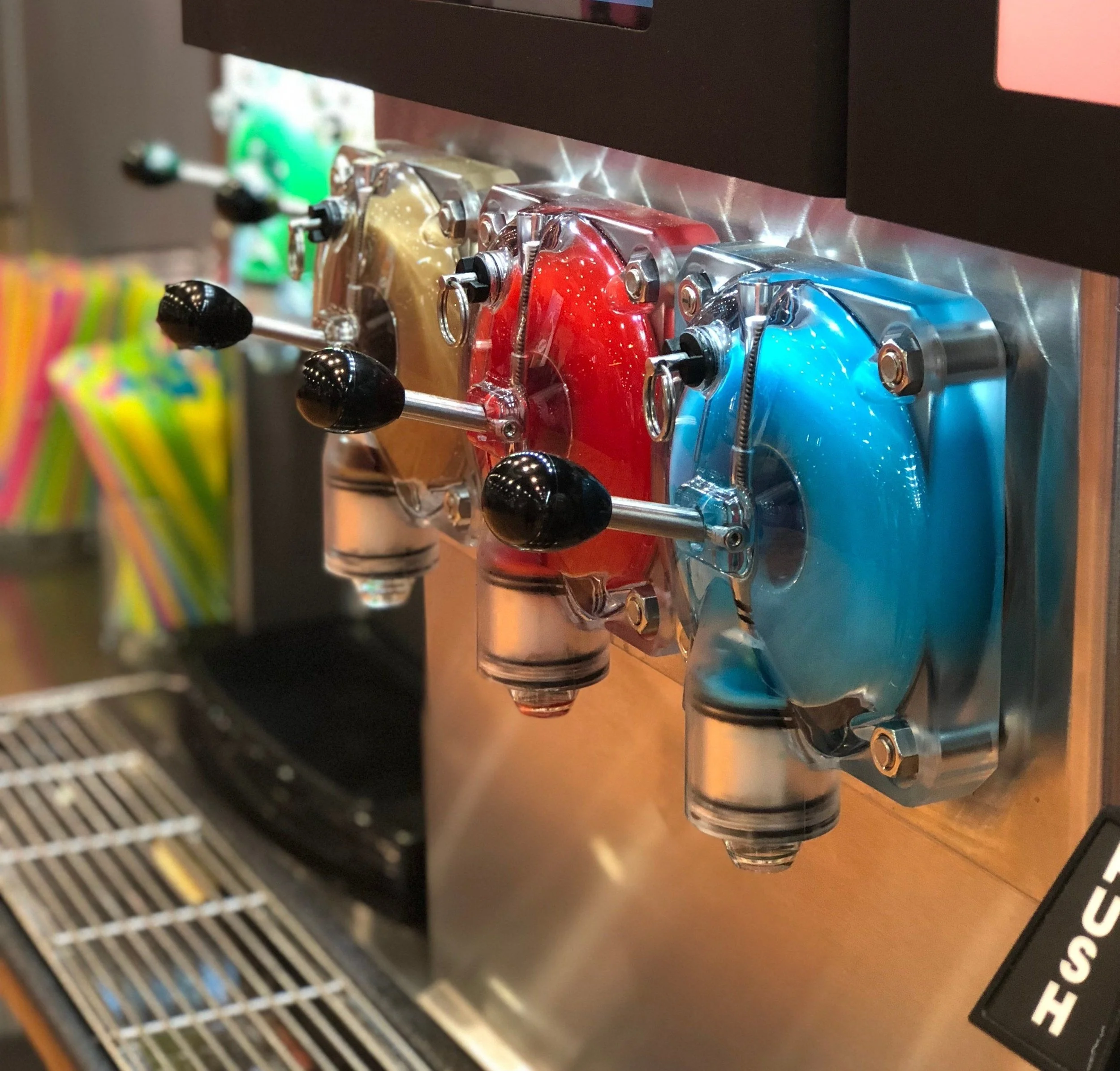 Three colorful soft-serve ice cream machines in red, blue, and gold, each with black handles, in a dessert shop.