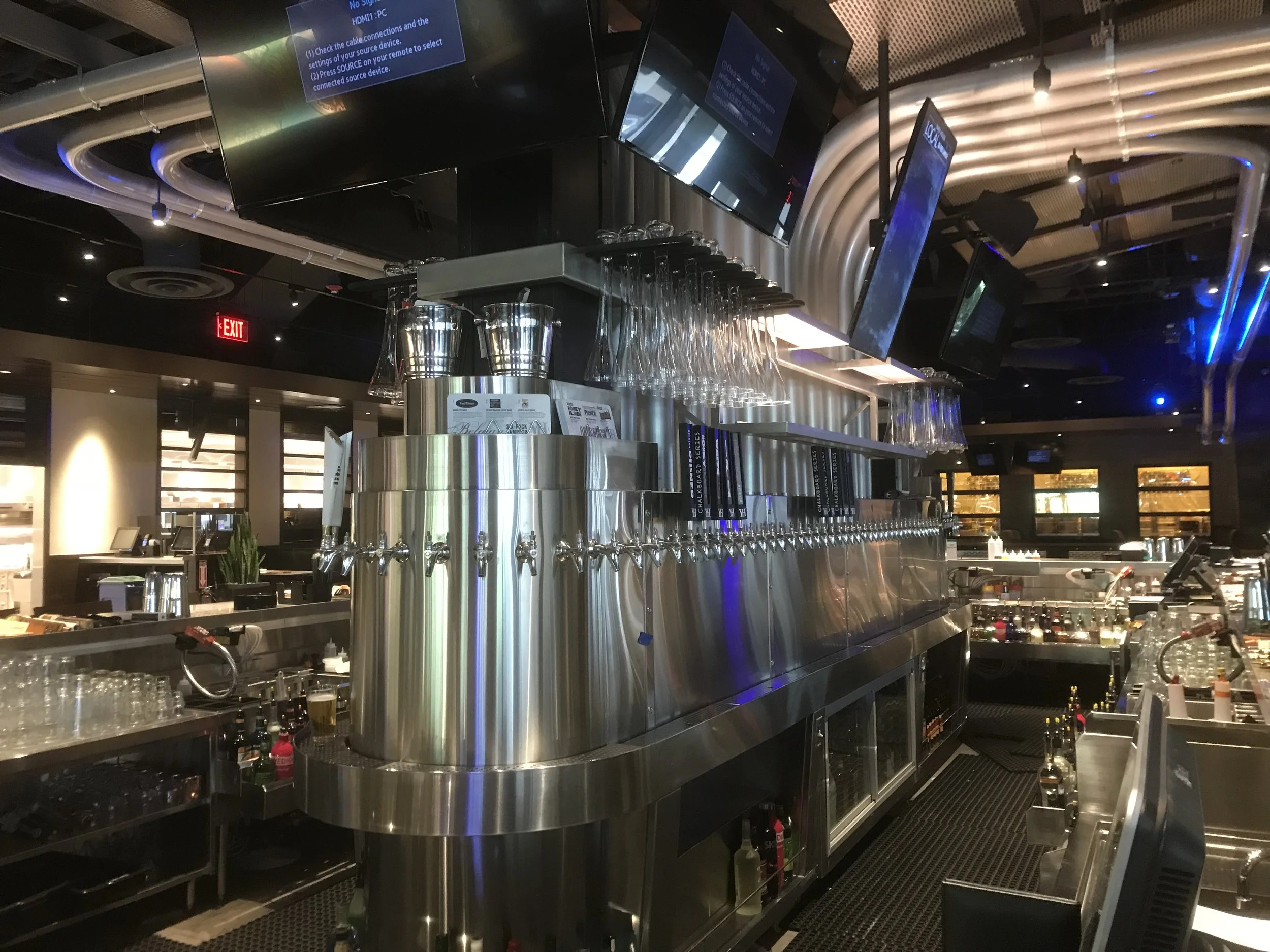 A stainless steel bar setup with multiple beer taps, glassware hanging above, and digital screens mounted on the ceiling in a modern bar or restaurant interior.