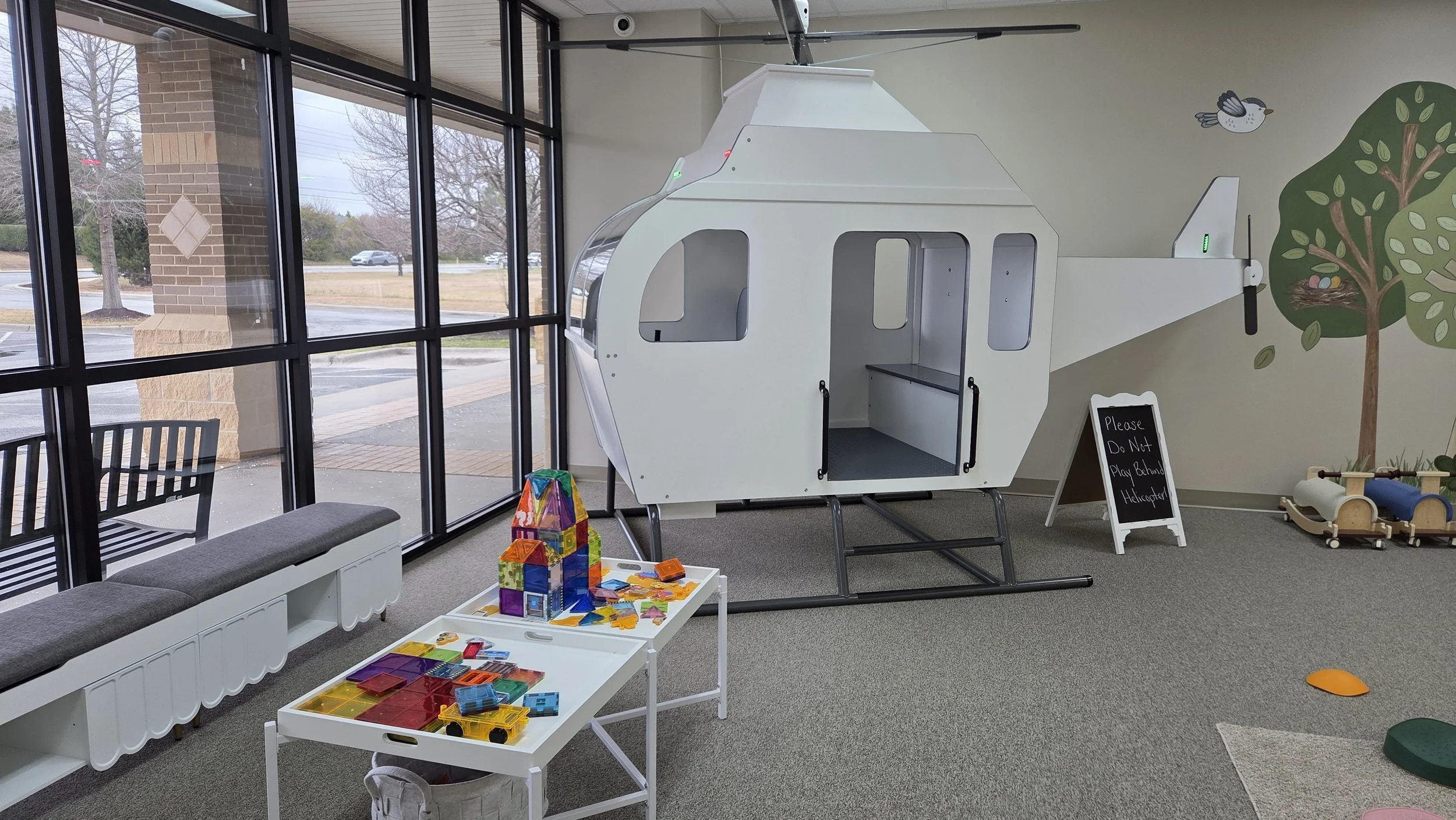 Kids play area with a small white helicopter-shaped play structure indoors near large windows, with colorful toys on white tables and a blackboard sign that says 'Please Do Not Play Behind Helicopter'.